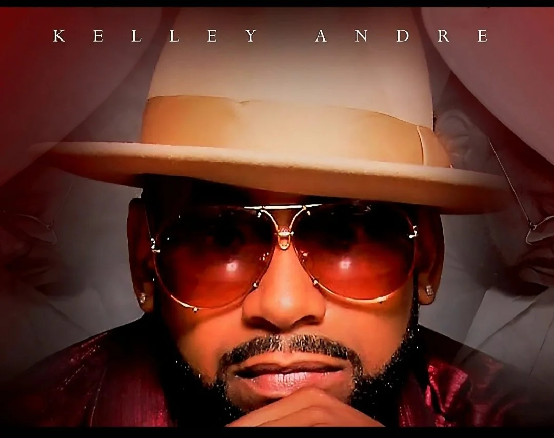Close-up of a man wearing large sunglasses, a beige hat, and earrings, with his hand resting on his chin, with the text "Kelley Andre" above his head.