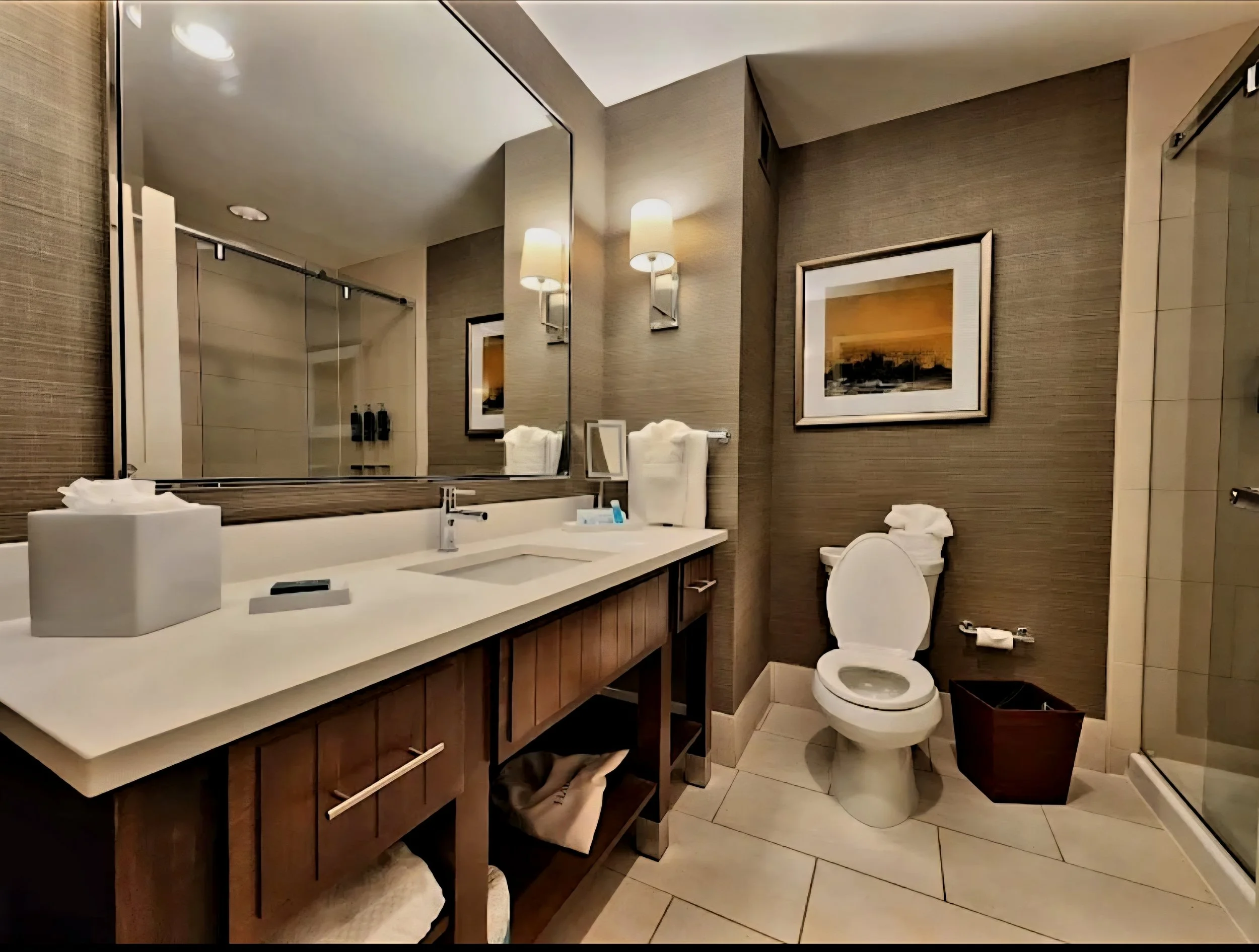 A modern hotel bathroom with a large mirror, two wall-mounted lights, a framed picture, a white sink with a faucet, neatly folded towels, and a toilet. There are toiletries on the counter and a trash bin on the floor.