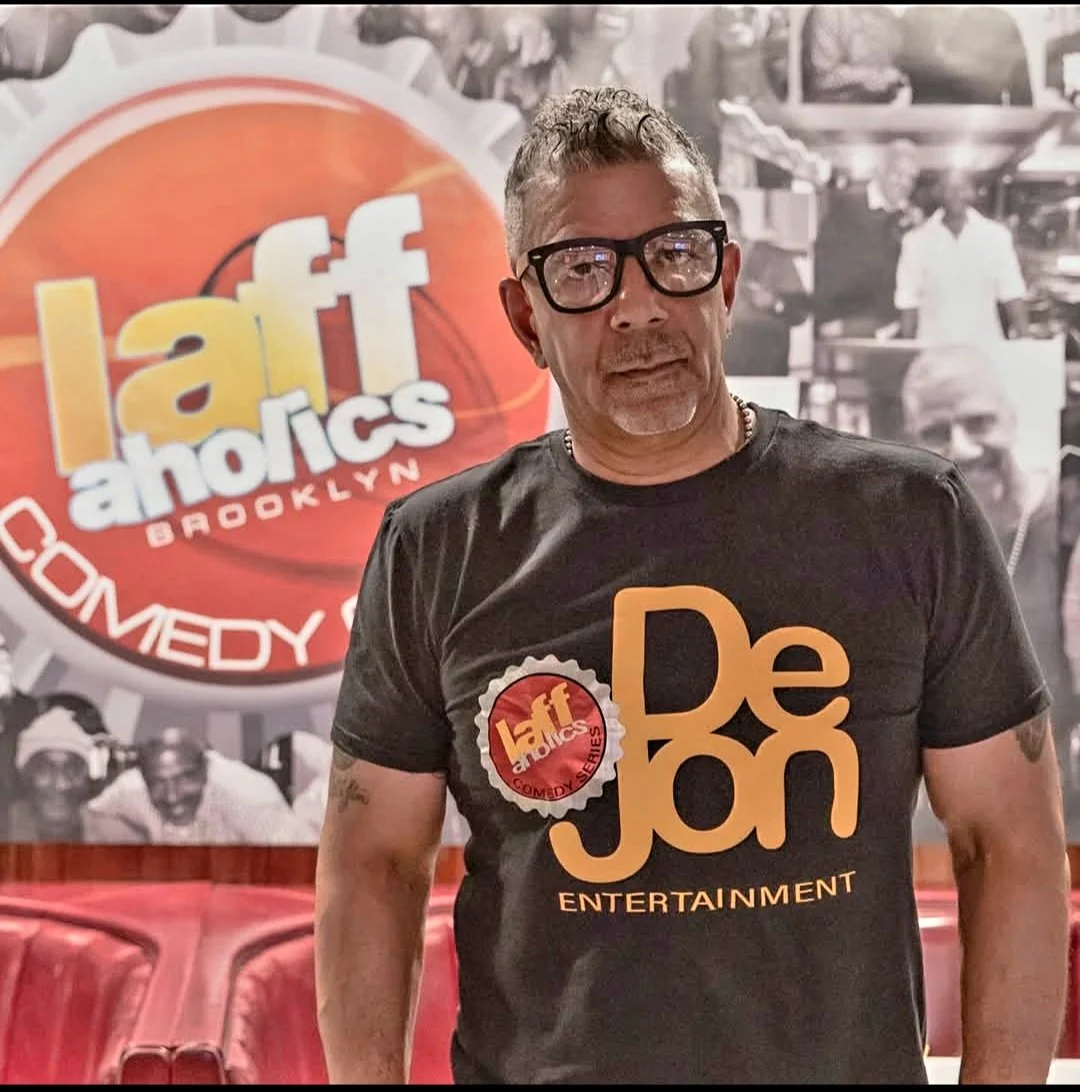 A man wearing glasses and a black T-shirt with the logo of Levity Brooklyn Comedy. The background features a large Levity Brooklyn logo and a black-and-white photo of a busy market scene.