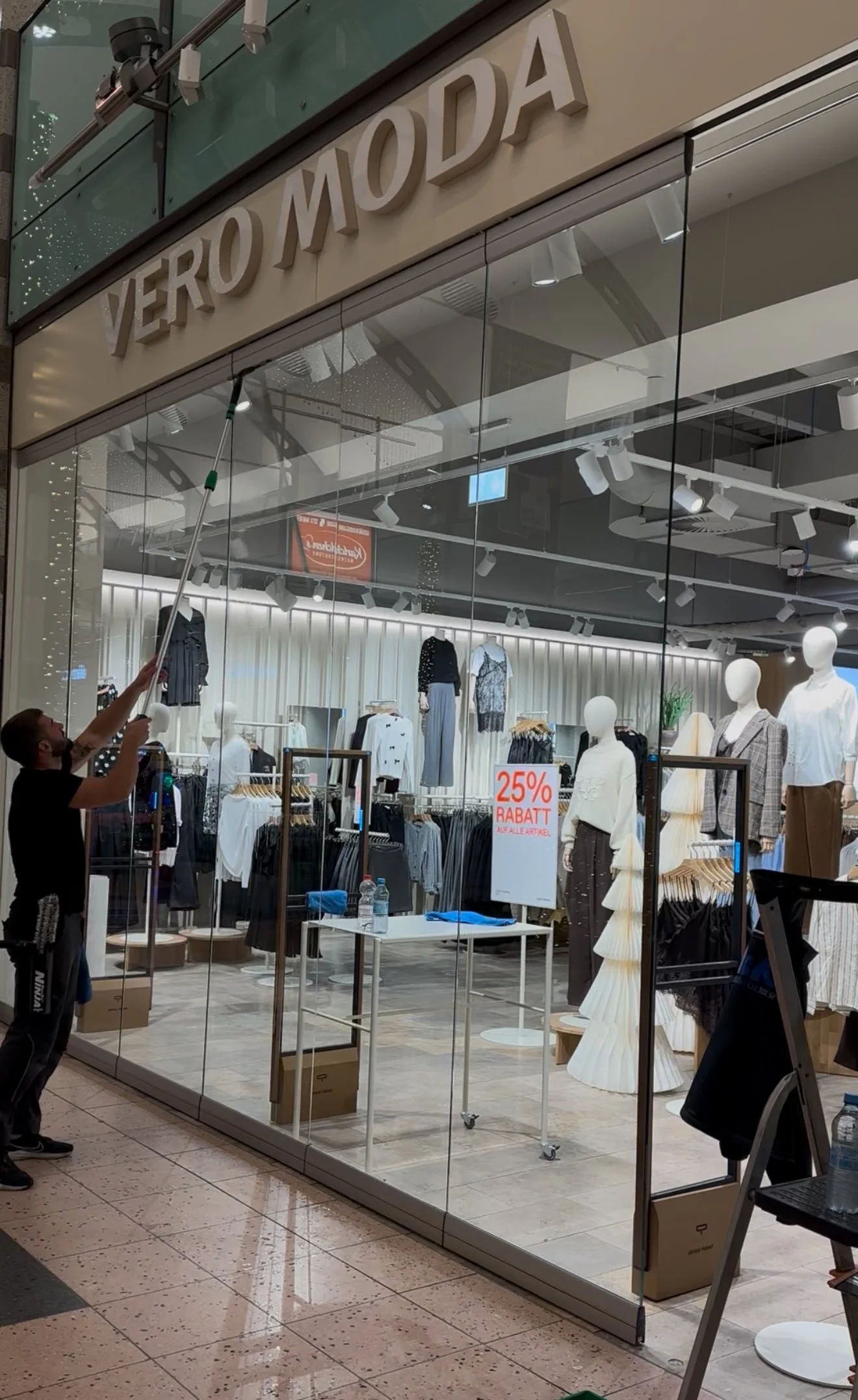 Mann reinigt die Schaufensterfront eines Modegeschäfts namens VERO MODA, das Kleidung und Accessoires verkauft. Es gibt einen Aufkleber mit 25% Rabatt auf Auslaufartikel im Schaufenster.