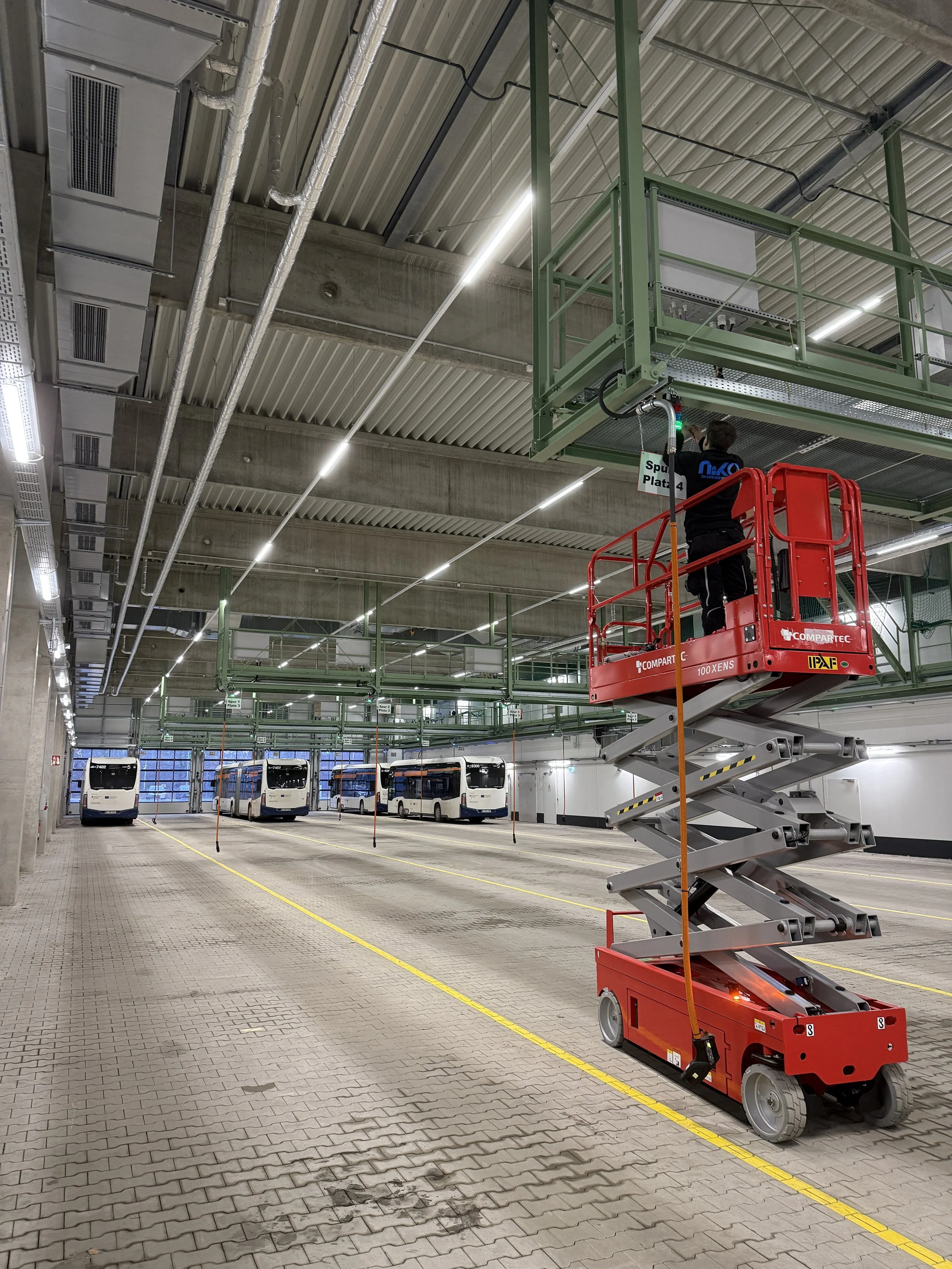 Ein Techniker arbeitet an der Decke eines Parkhauses mit mehreren parkenden Bussen. Er steht auf einem rot-weißen Hebewagen und verwendet ein Werkzeug an einer grünen Metallstruktur.
