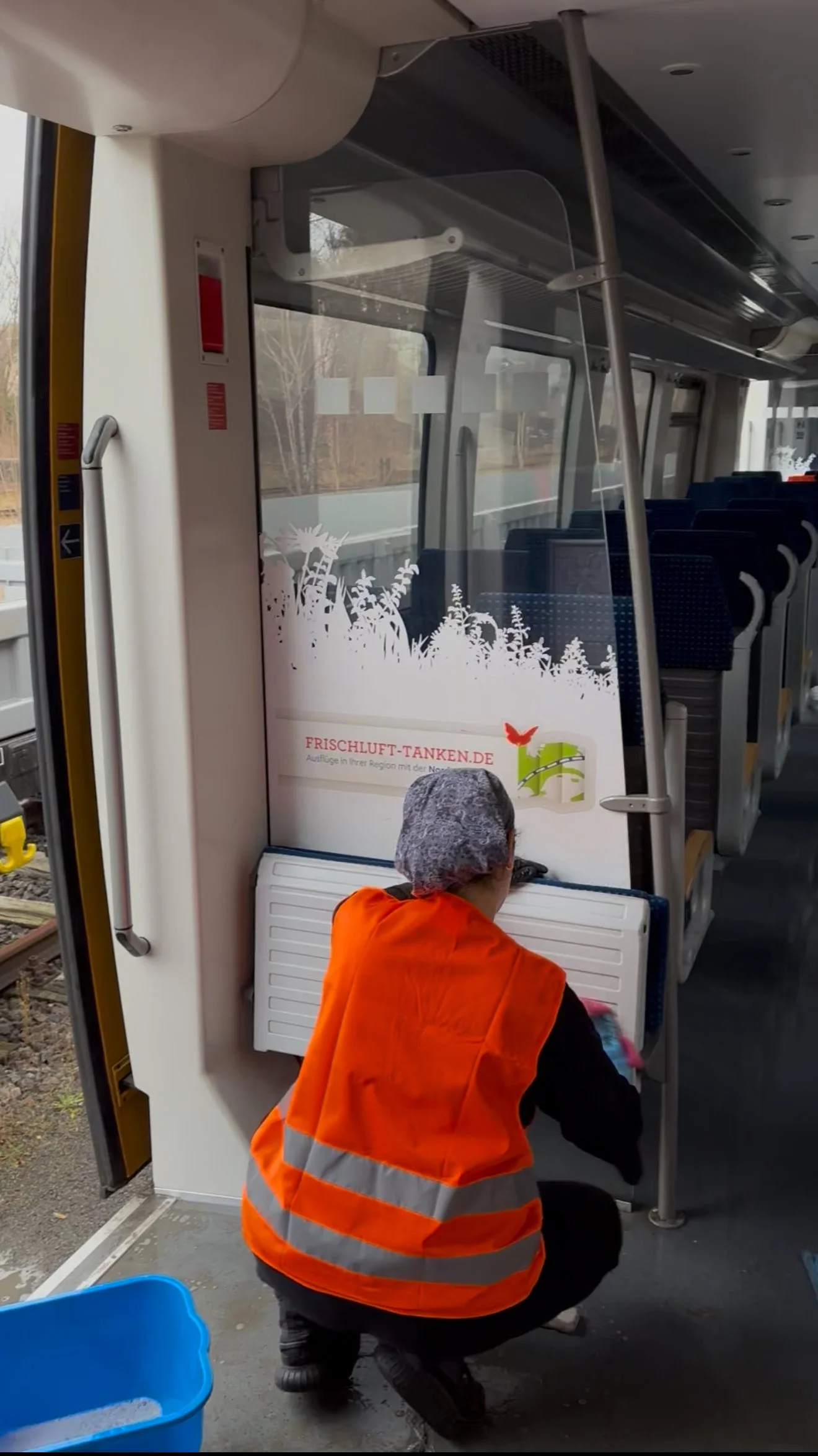 Eine Person in einer orangefarbenen Sicherheitsweste sitzt in einem Bus, die Person hat eine Haube auf und scheint Wasser zu reinigen oder eine Aufgabe zu erledigen. Im Hintergrund sind Businnenraum-Sitze und eine Scheibe mit einer dekorativen Folie 