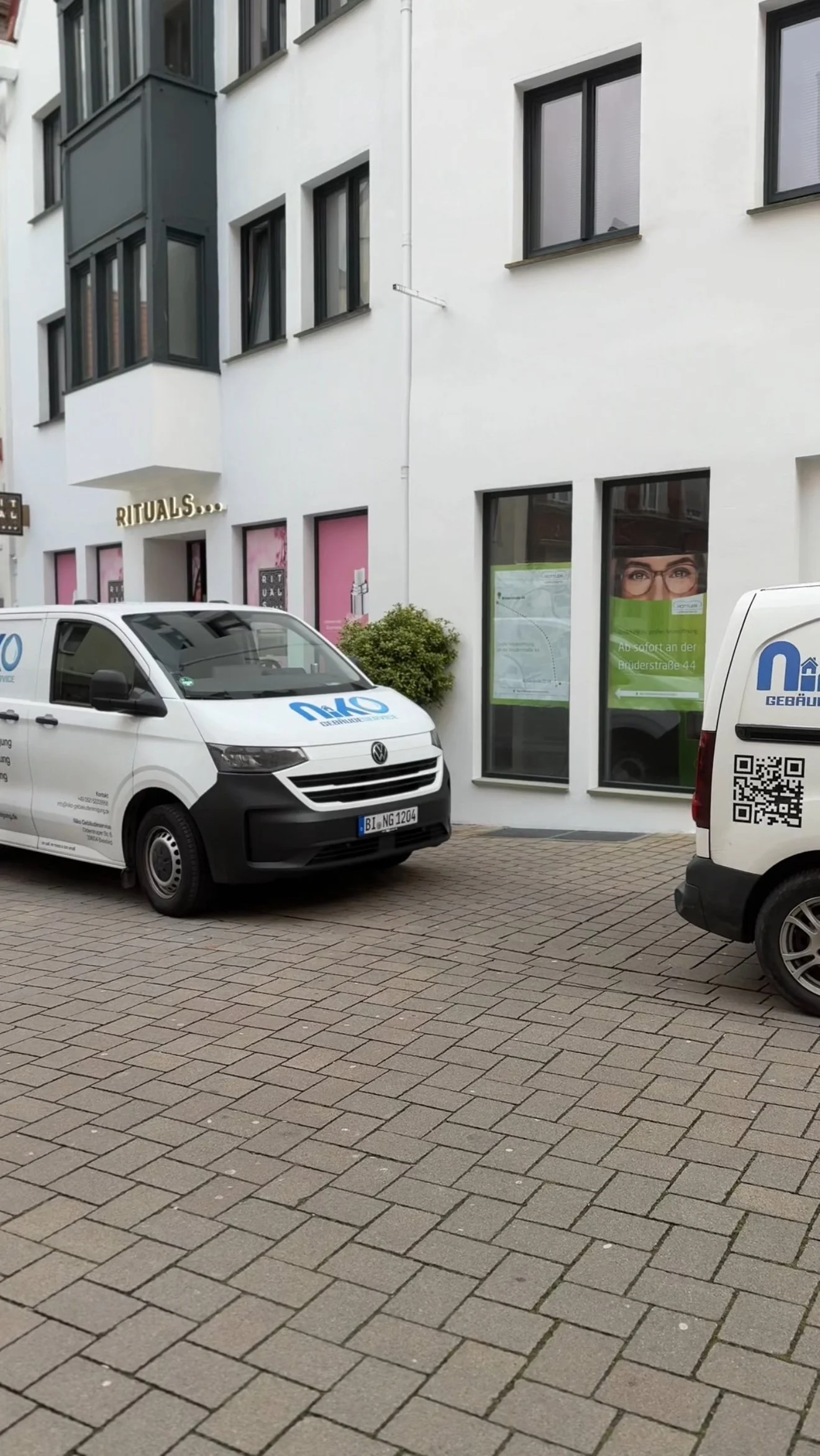 Zwei weiße Firmenwagen parken auf einem gepflasterten Straßenabschnitt vor einem weißen Gebäude mit mehreren Fenstern und einem Geschäft namens 'Rituals'.