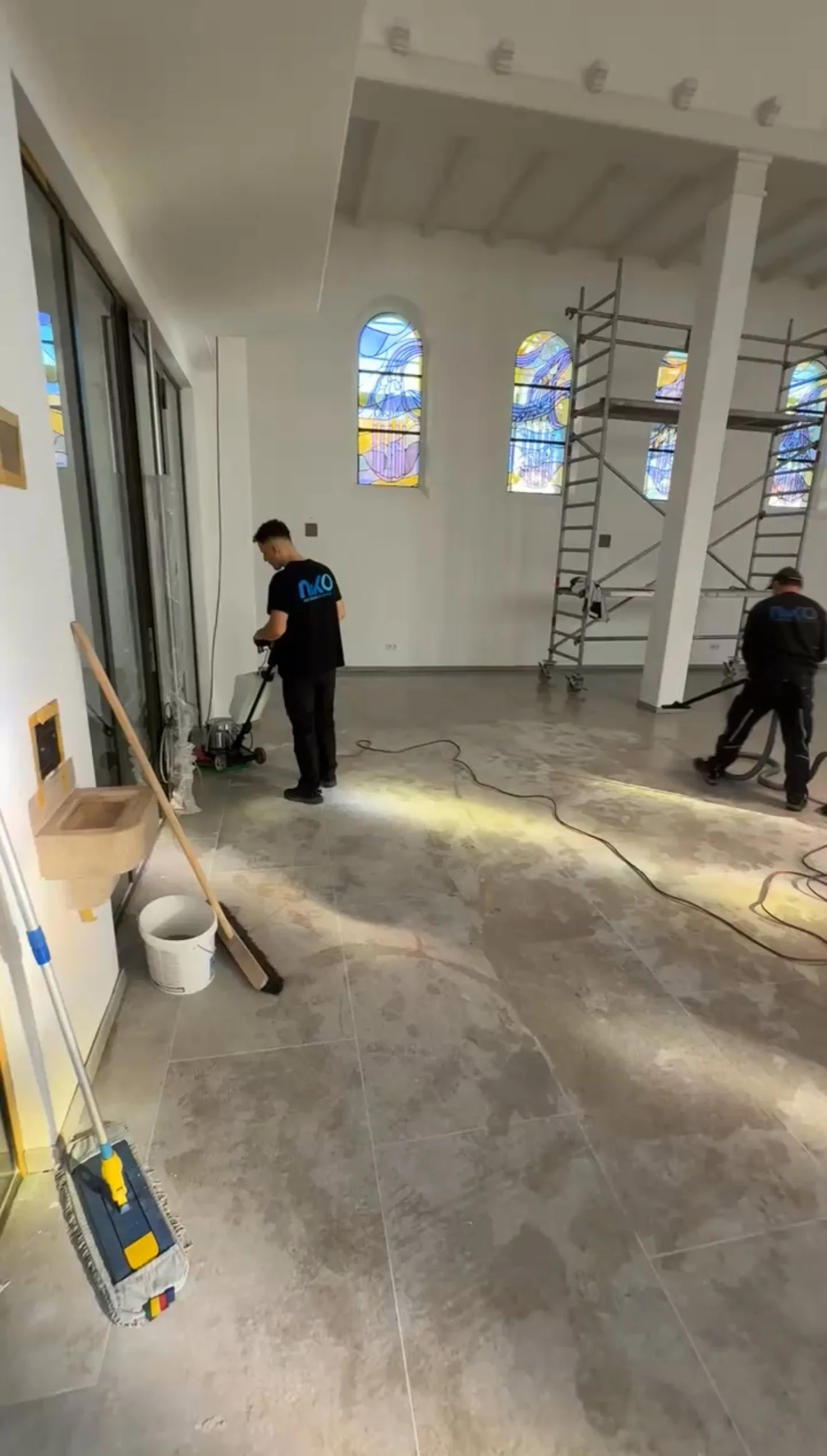 Zwei Arbeiter reinigen den Boden in einem Raum mit bunten Kirchenfenstern; eine Person verwendet eine Schleifmaschine, die andere eine Maschine zur Staubreinigung; im Raum sind Gerüste aufgebaut.