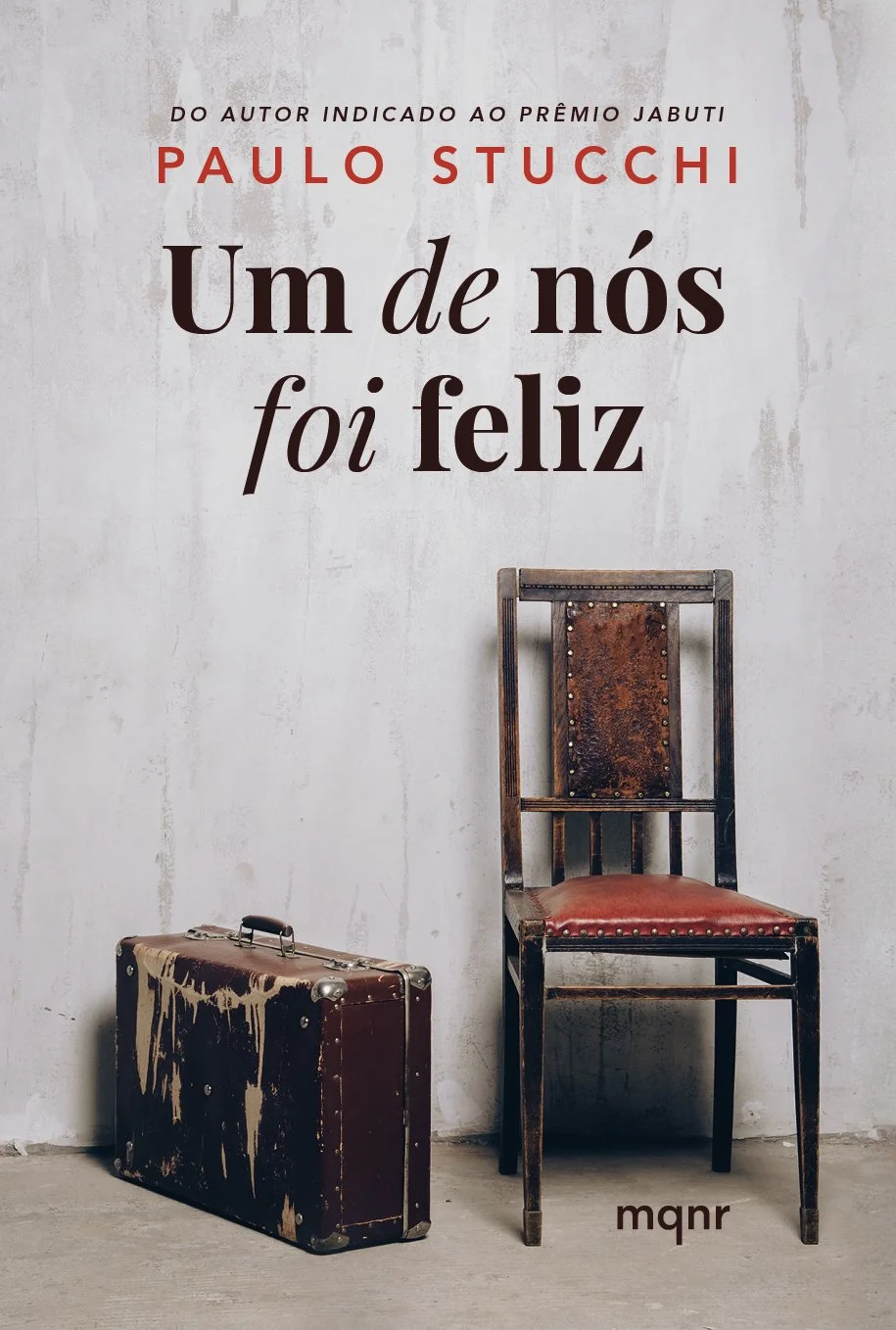 Capa de livro com uma cadeira de madeira envelhecida, uma mala antiga suja ao lado, e o título "Um de nós foi feliz" de Paulo Stucci em uma parede de fundo clara.