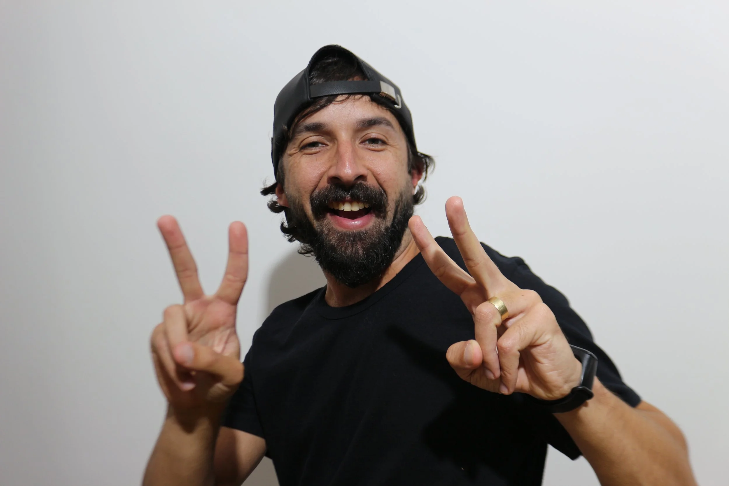 Homem sorridente e feliz, usando boné para trás, fazendo o gesto da paz com as duas mãos, com fundo branco.