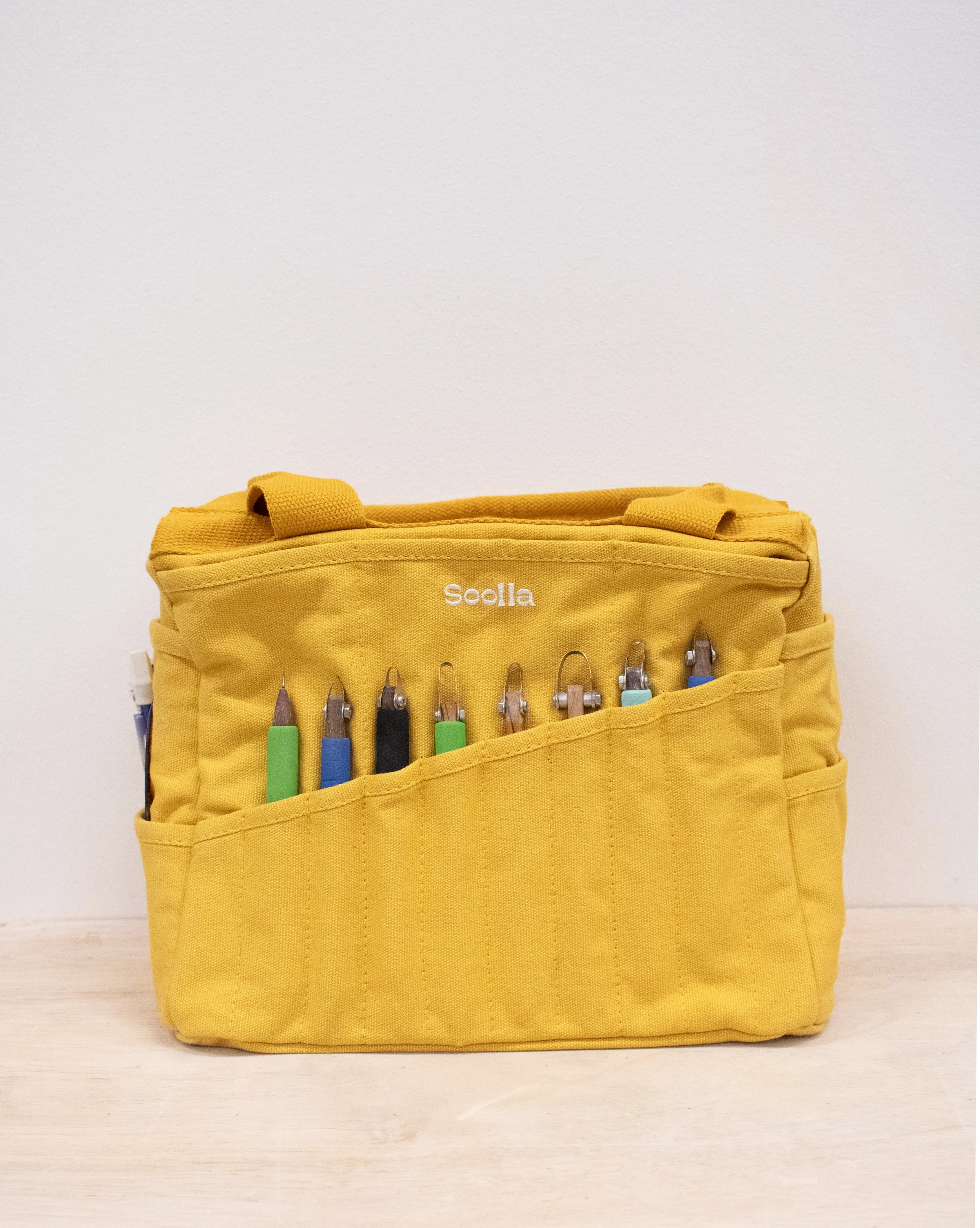 Yellow Soolla brand tool bag with various crochet hooks stored in the front pocket, placed on a light wooden surface against a white wall.