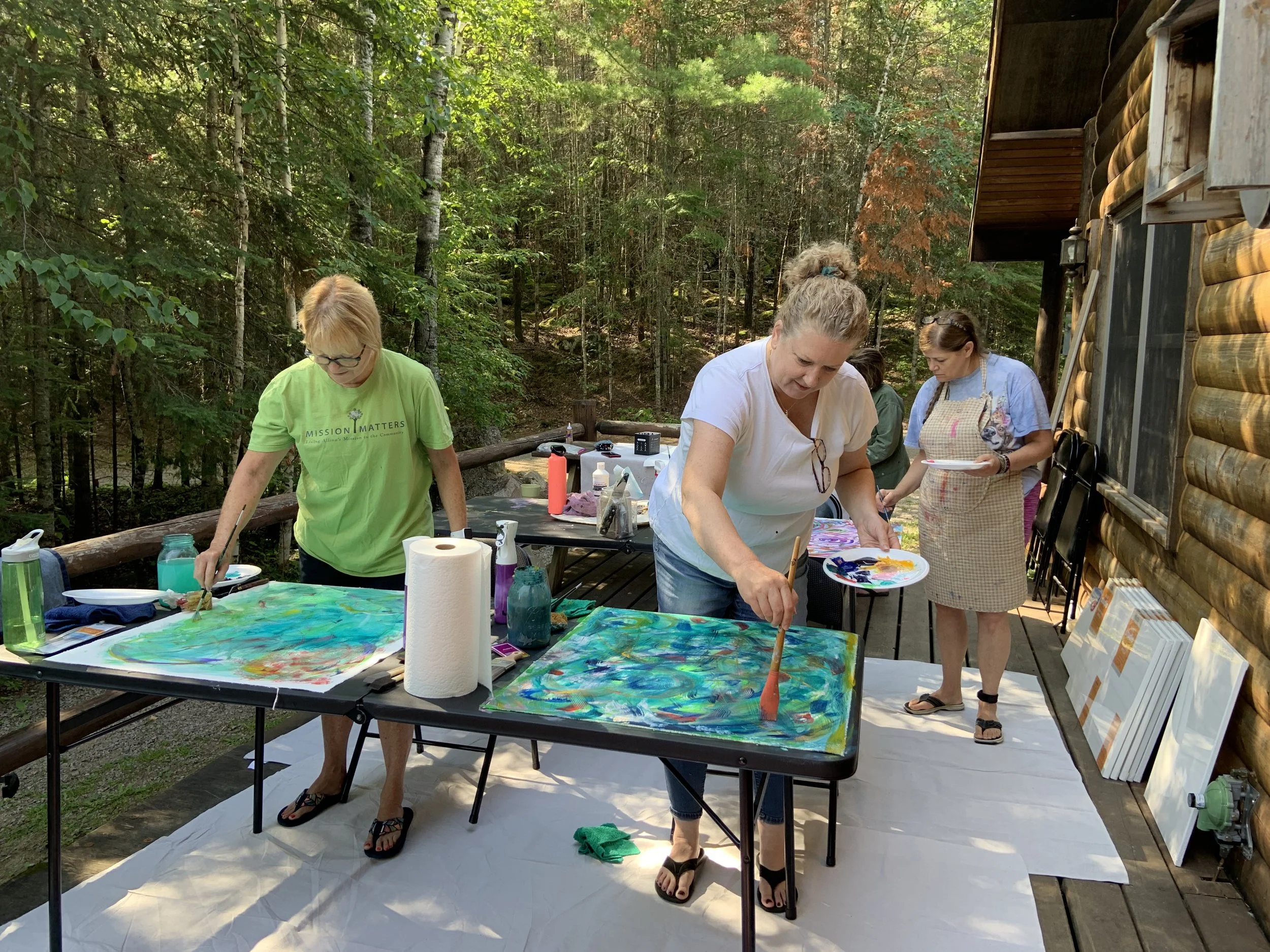Three women painting canvases on a deck surrounded by trees and a log cabin wall.