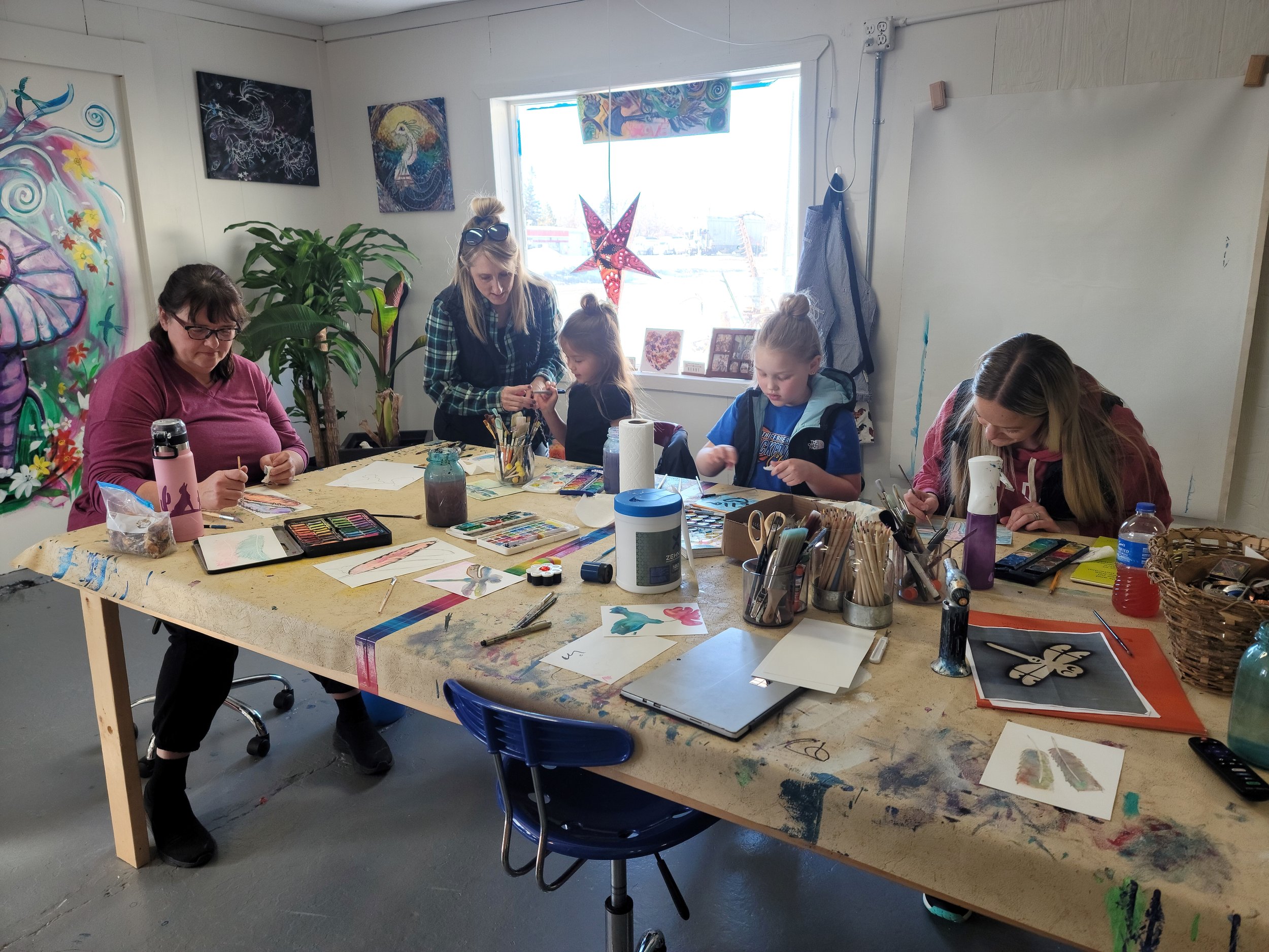 People crafting at a wooden table in an art studio with colorful paintings on the walls.