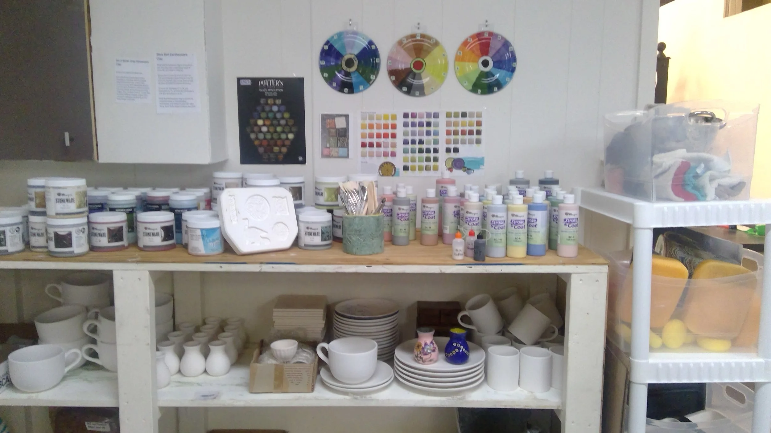 Pottery studio supplies including paint jars, color charts, brushes, and ceramic pieces on shelves.