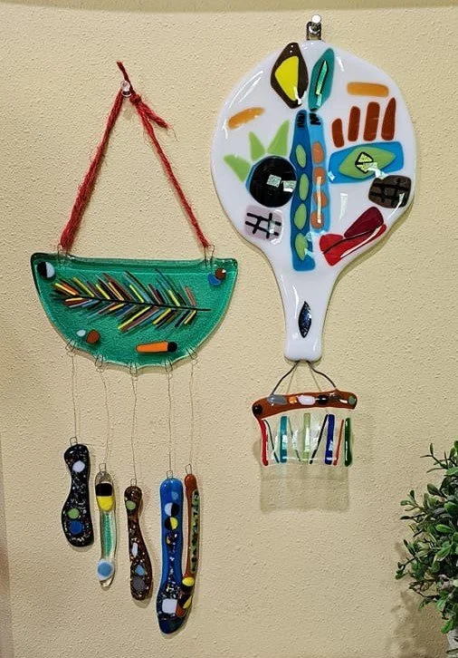 Colorful fused glass art pieces hanging on a wall, featuring abstract designs with various geometric shapes and vibrant colors, connected by strings.