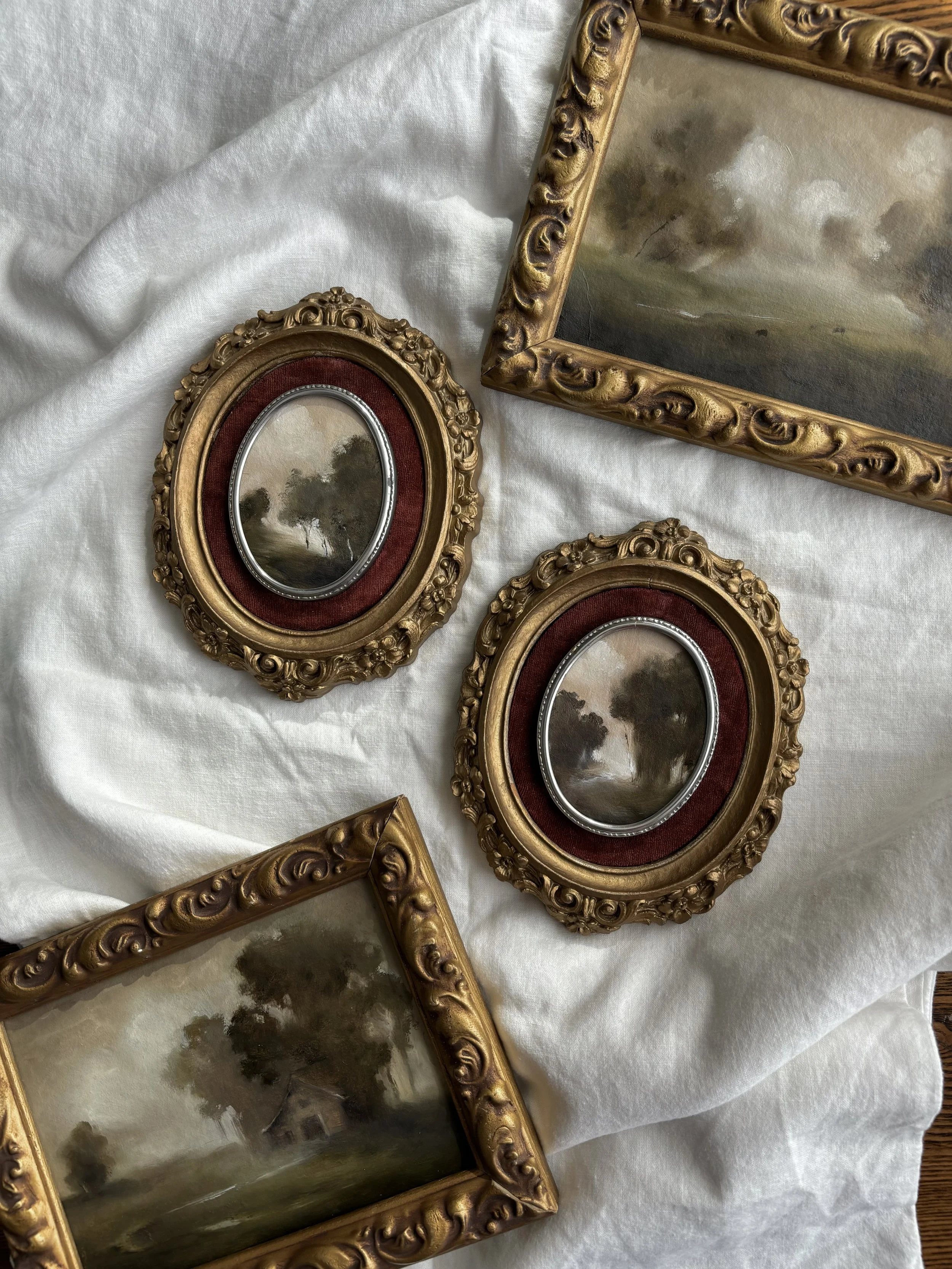 Collection of small landscape paintings in ornate gold frames on white fabric background.