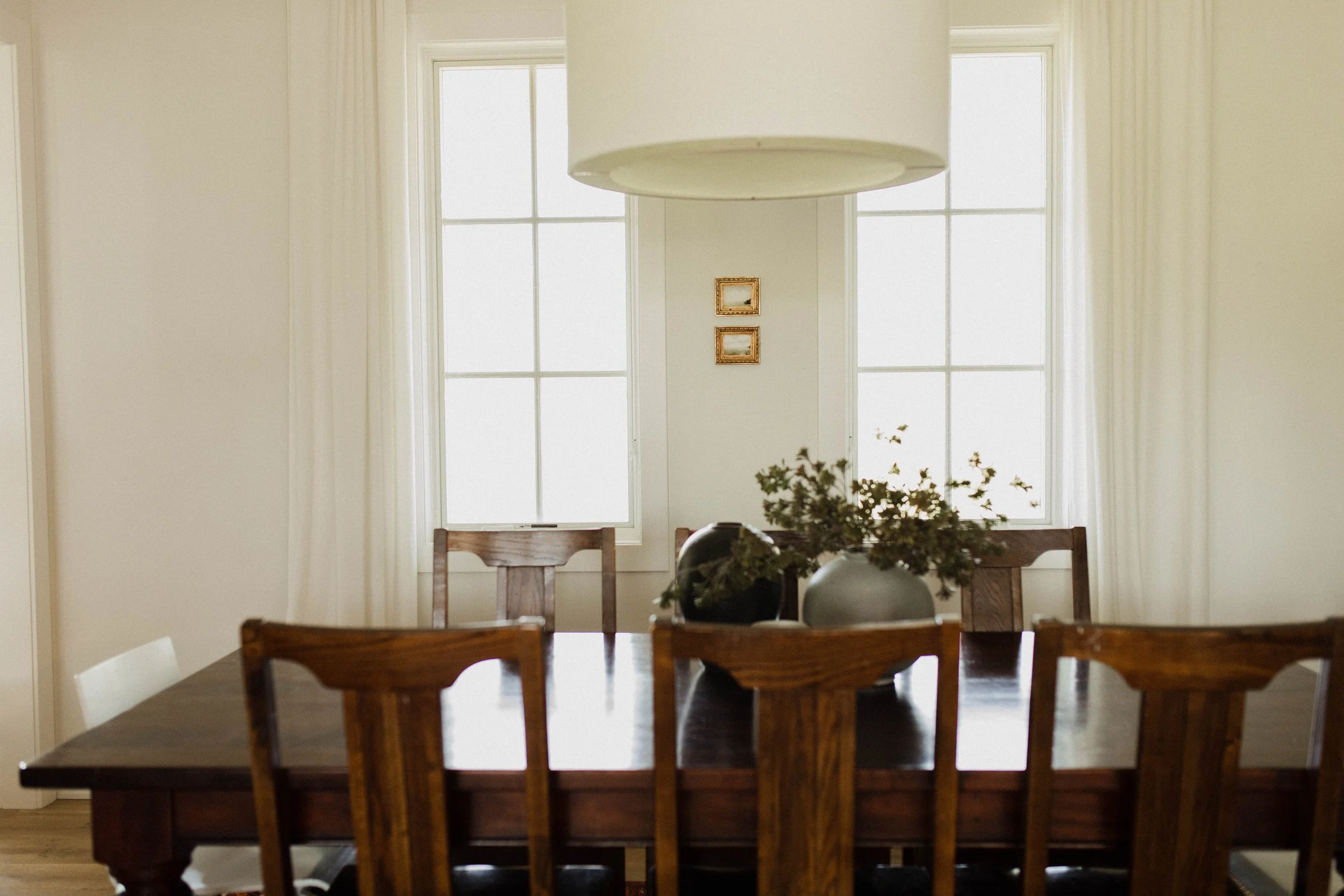 Dining room with wooden table, chairs, green foliage centerpiece, pendant light, and framed pictures on light-colored walls.