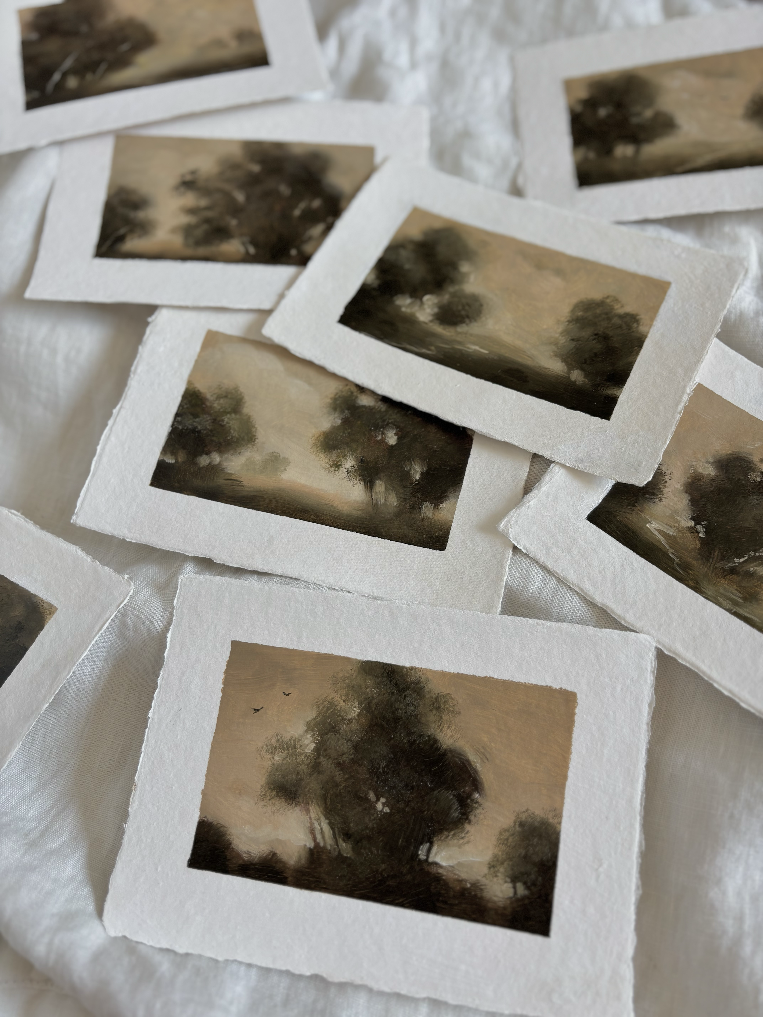 Several small landscape paintings of trees and cloudy skies, arranged on a white cloth surface.