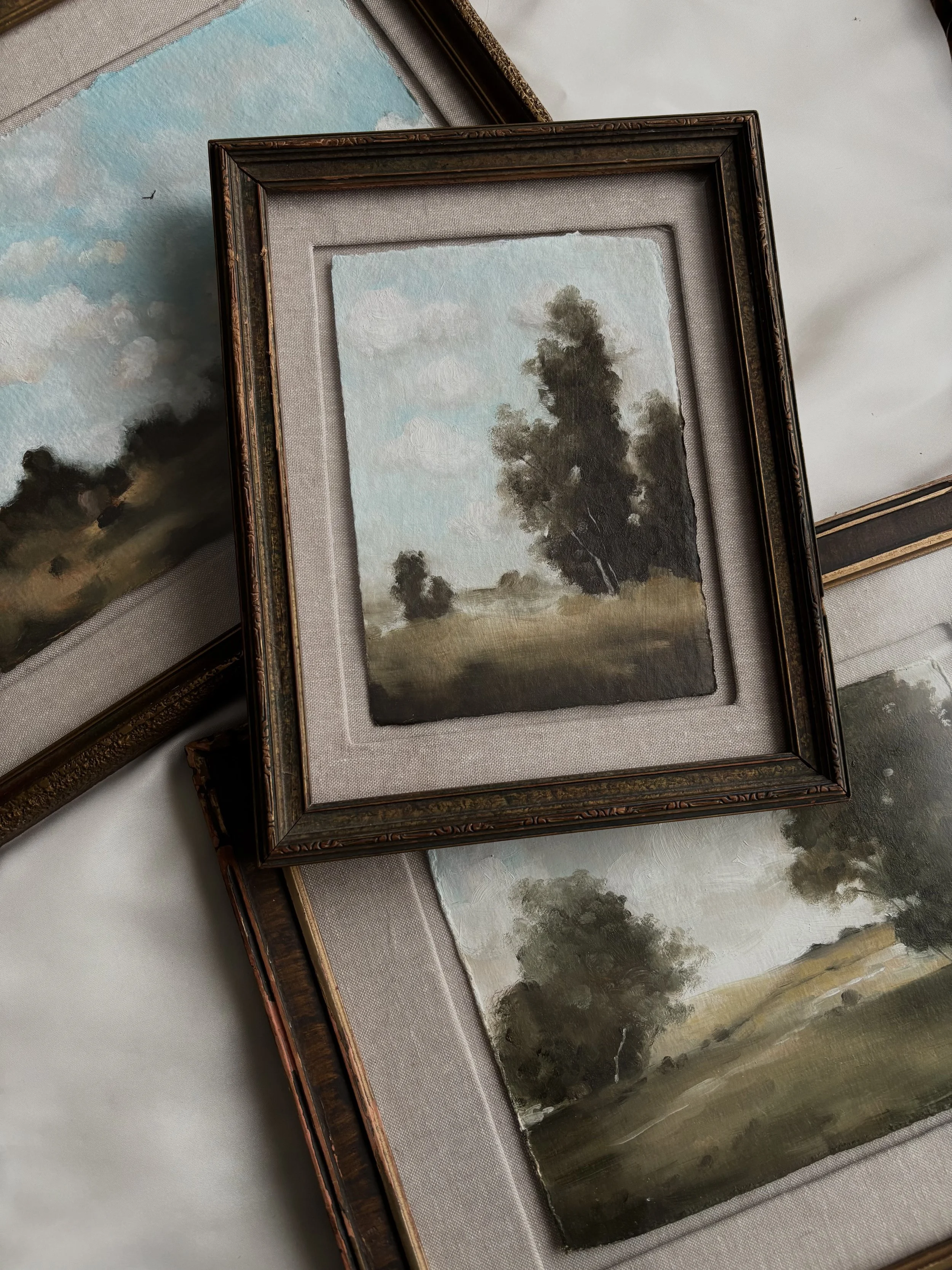 Three framed landscape paintings with trees and fields, featuring pastel skies and grass. The frames are rustic and antique-style.