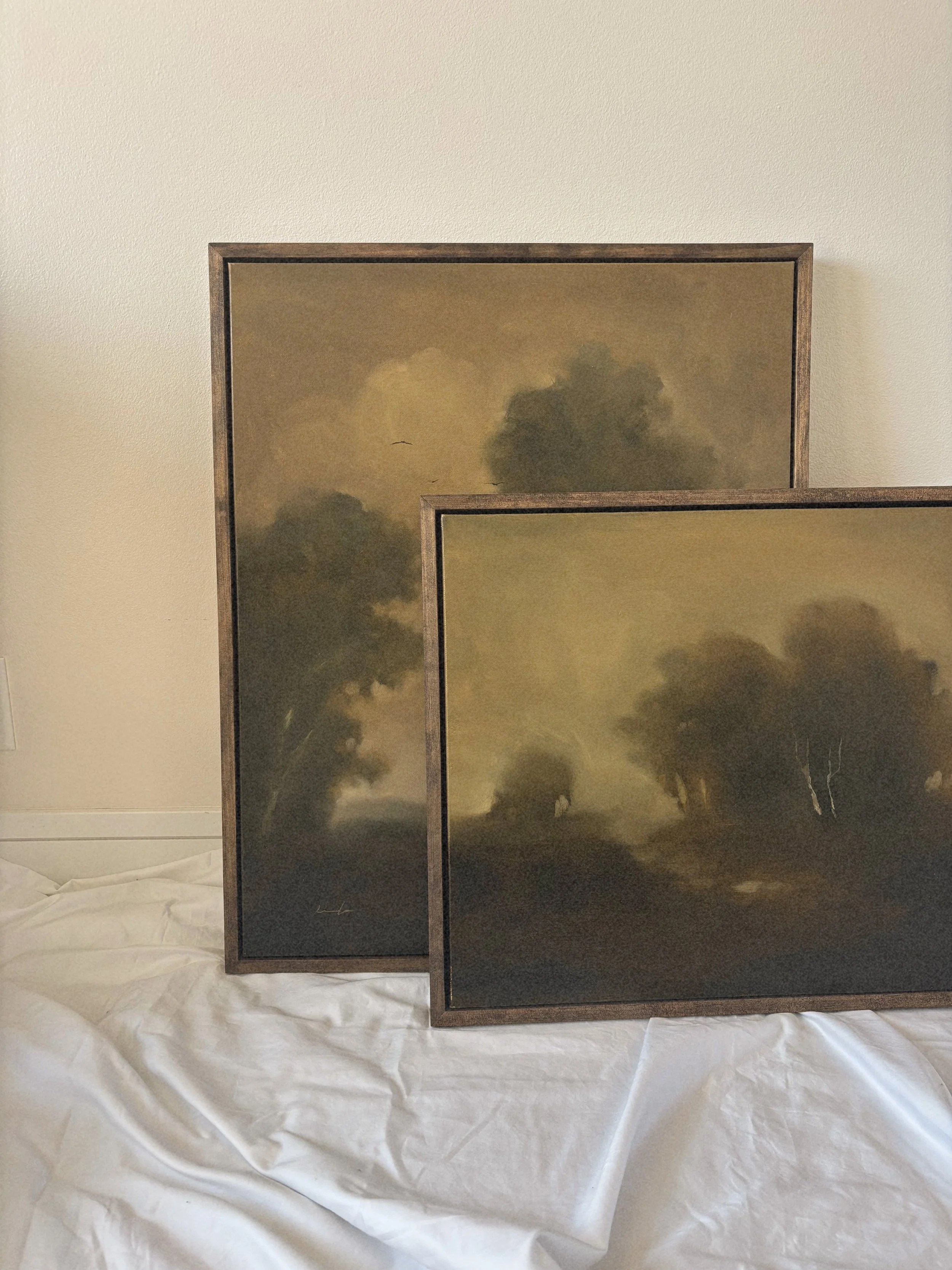 Two framed landscape paintings with cloudy sky scenery, placed on a crumpled white surface, against a light-colored wall.
