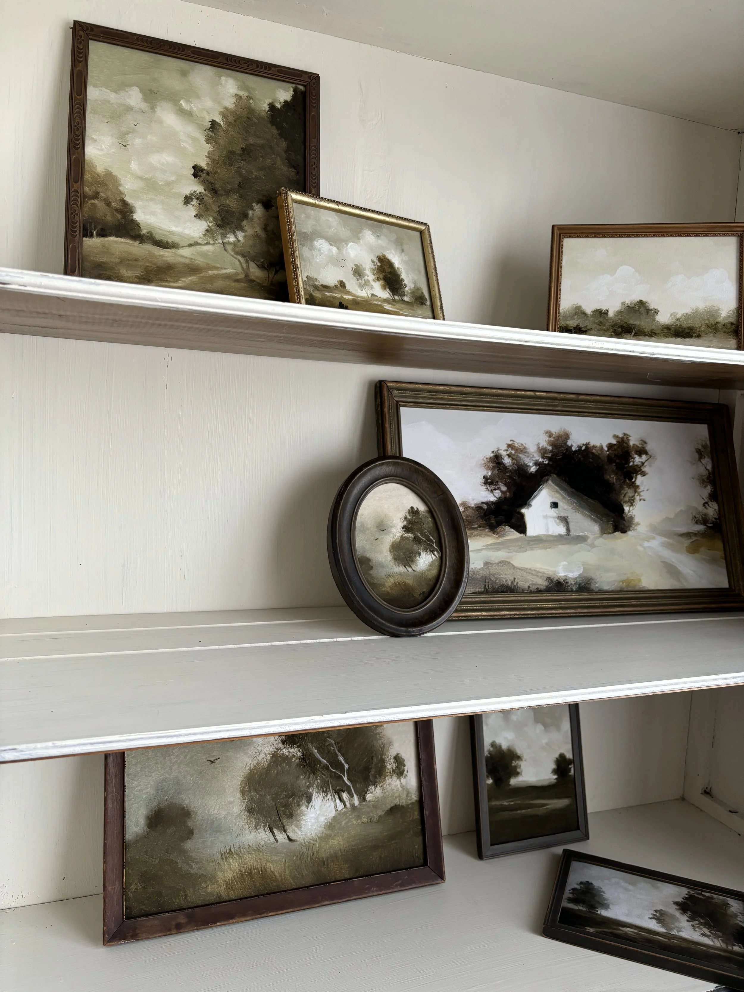 Assortment of framed landscape paintings on white shelves, featuring trees and countryside scenes, mostly in sepia tones.