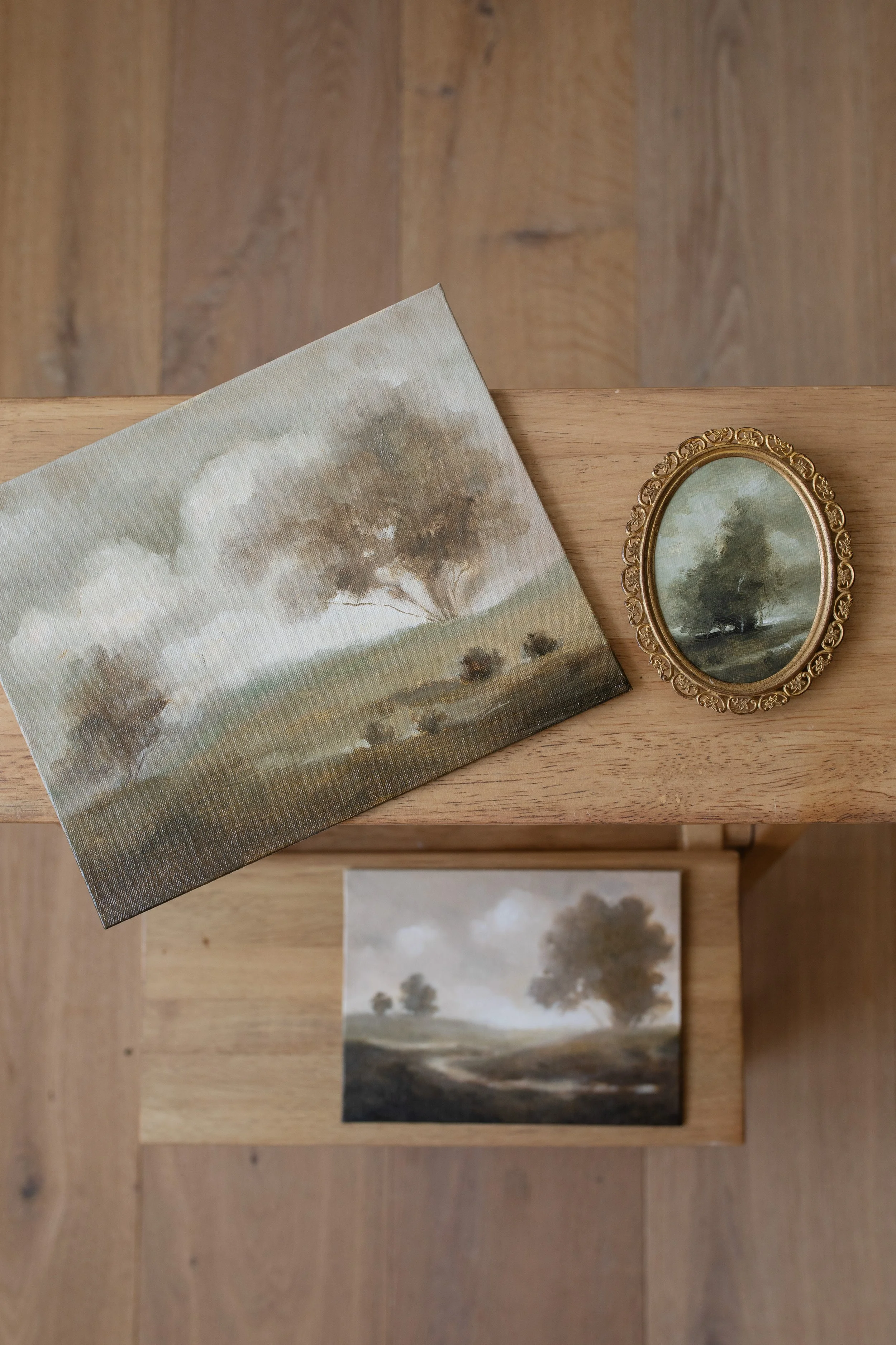 Three small landscape paintings in earthy tones, displayed on wooden surfaces, with one in an ornate oval frame.