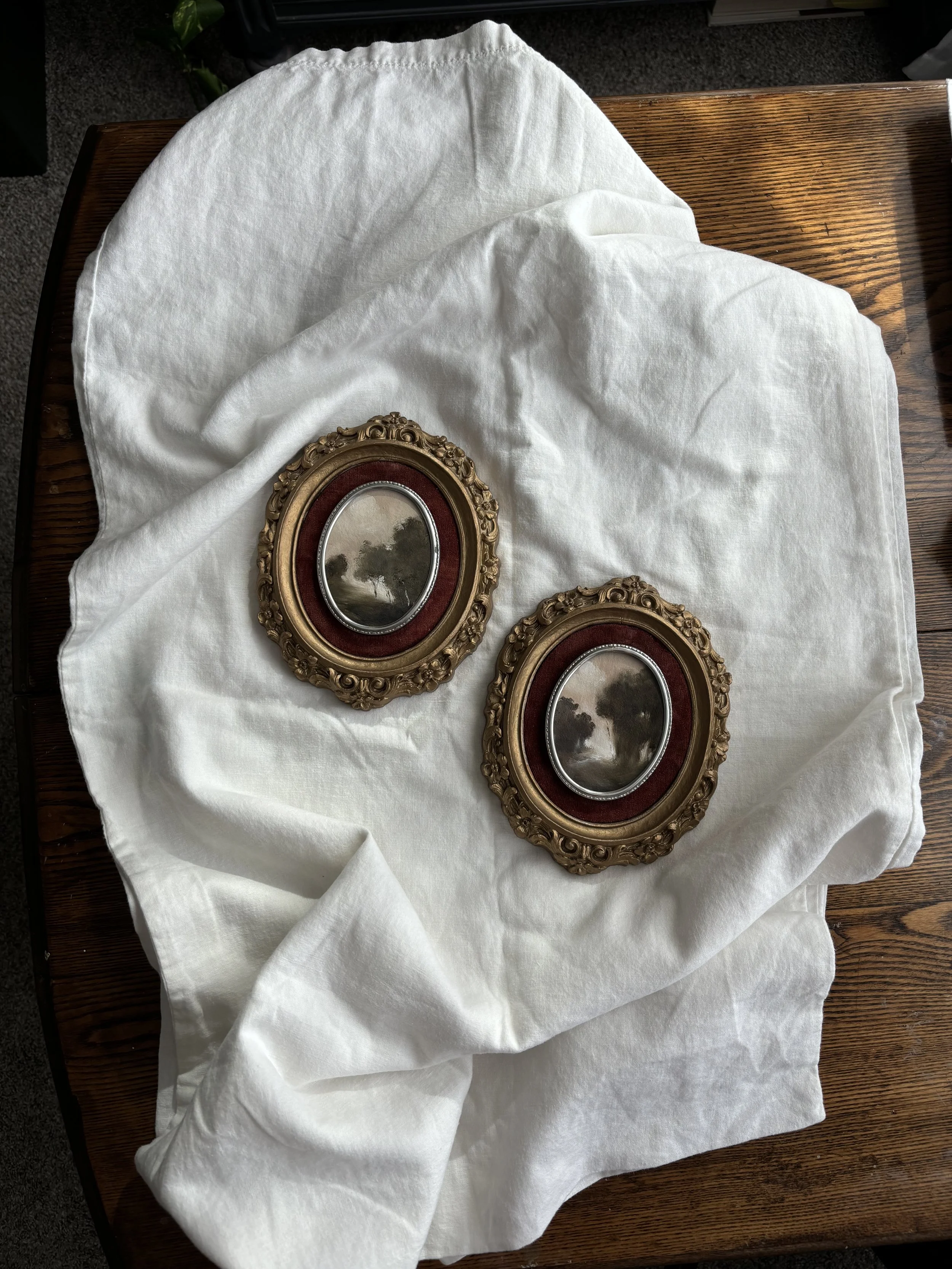 Two vintage-style oval photo frames with ornate gold detailing, placed on a white cloth on a wooden surface.