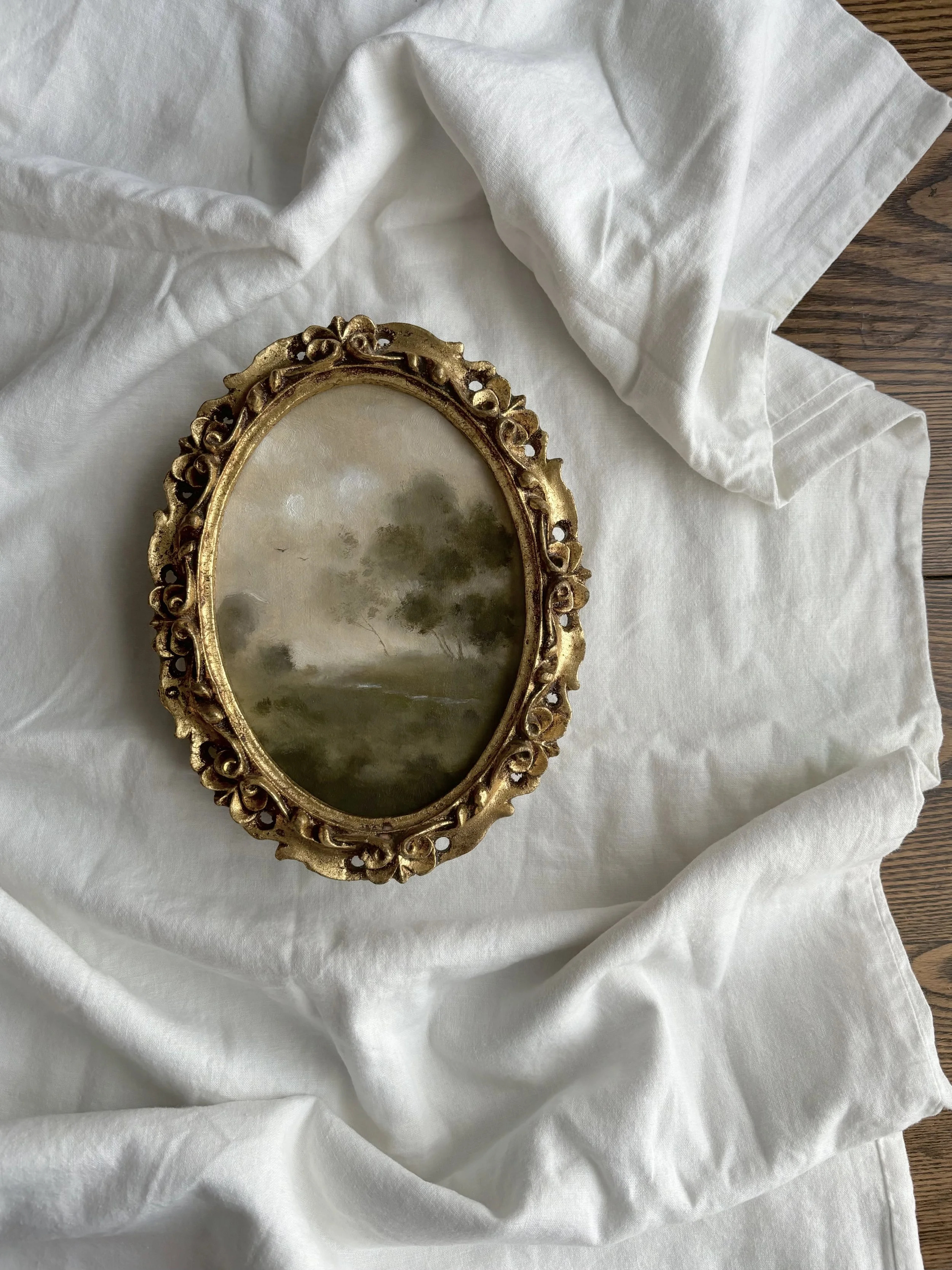 Vintage oval picture frame with gilded floral design on white fabric background.