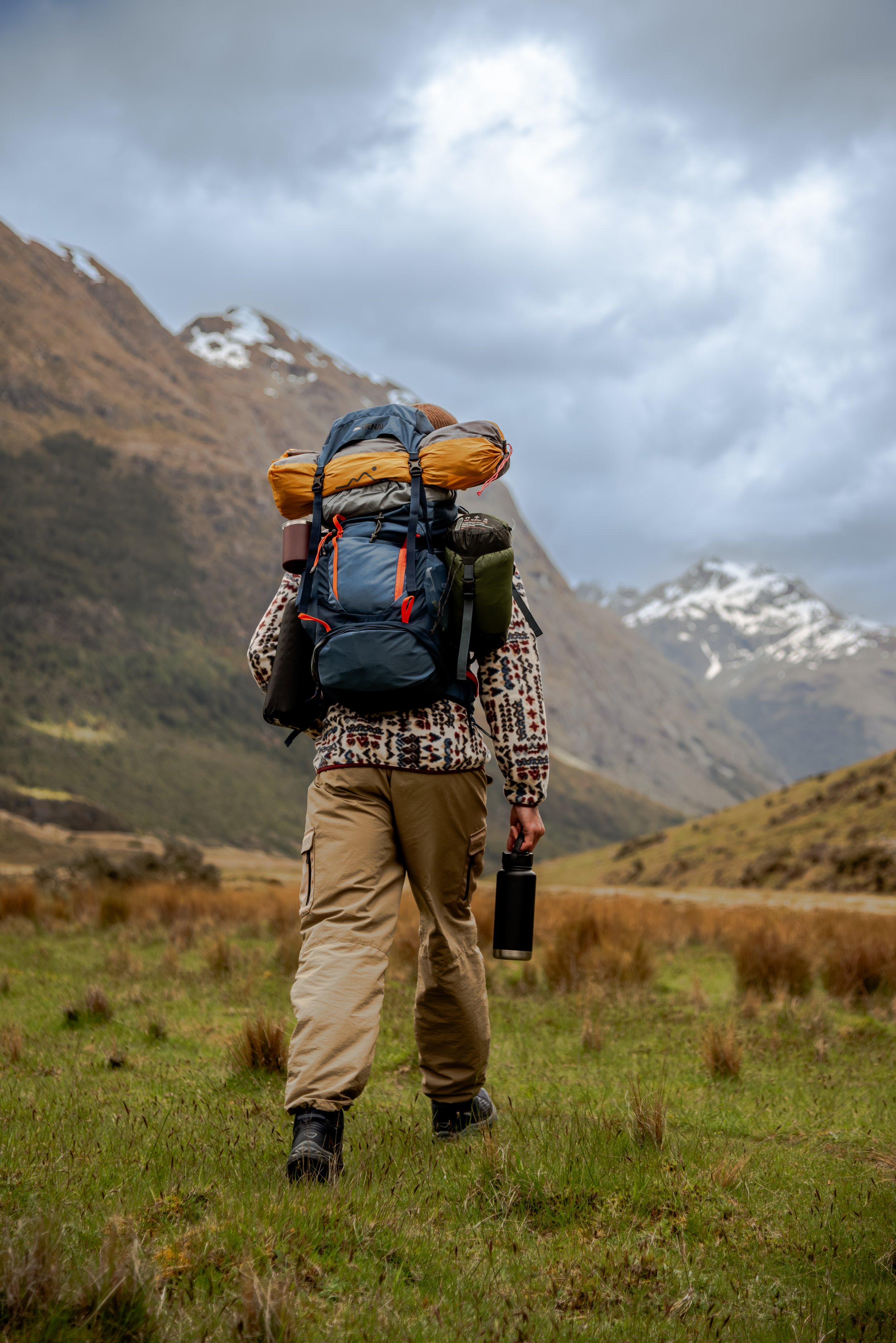 A hiker with a large backpack and water bottle walking through a grassy, mountainous landscape on a cloudy day.