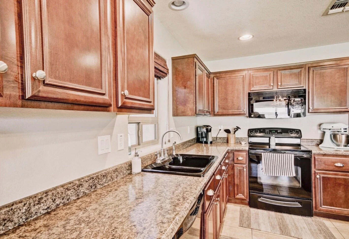 Kitchen with wooden cabinets, granite countertops, black oven and microwave, small window above the sink, and various kitchen appliances.