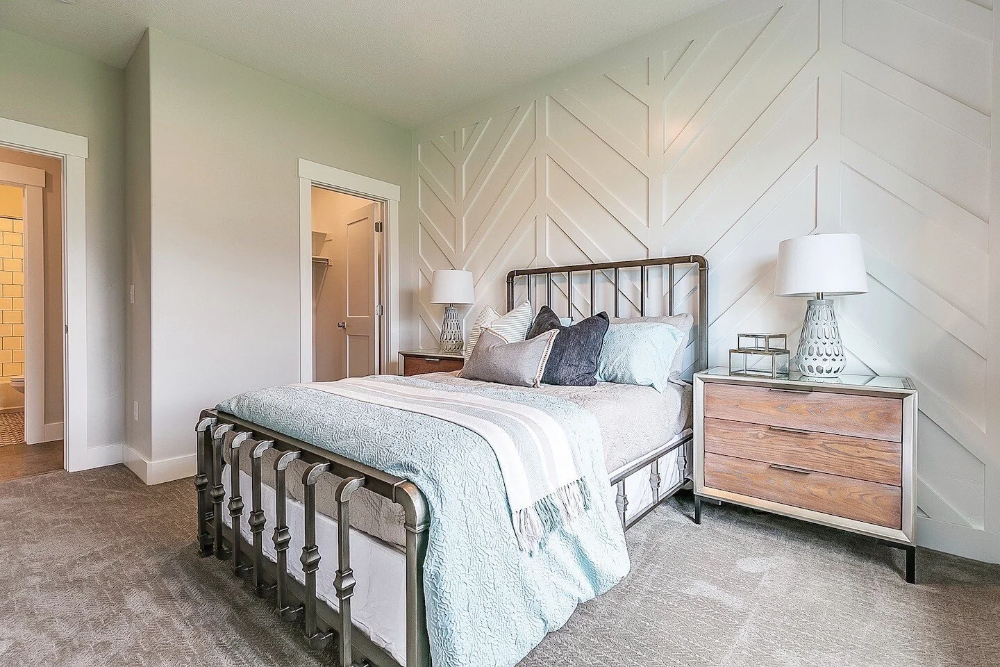 Modern bedroom with a metal bed frame, white bedding, and multiple decorative pillows, flanked by nightstands with table lamps, and a textured angled wall behind