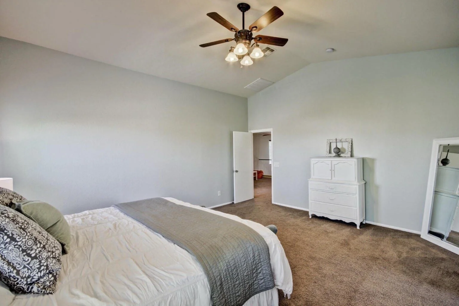 A bedroom with a bed on the left featuring decorative pillows and a gray blanket, a white dresser with a vase and framed mirror above it on the right, a large mirror leaning against the wall, brown carpeted flooring, a central ceiling fan with lights