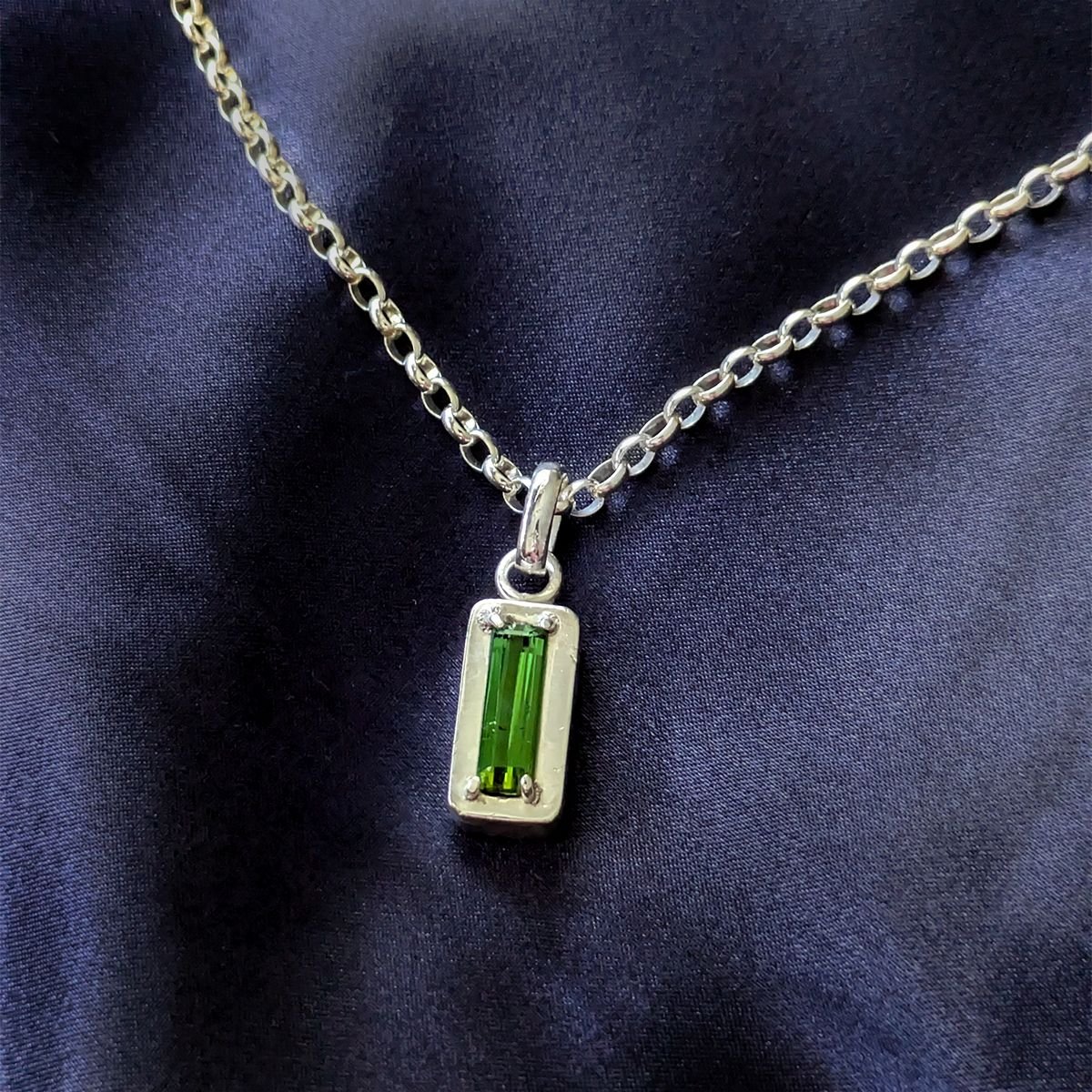 Close-up of the Ohad necklace with a baguette-cut green tourmaline claw-set on a fine silver platform, on a sterling silver Belcher chain over dark navy fabric.