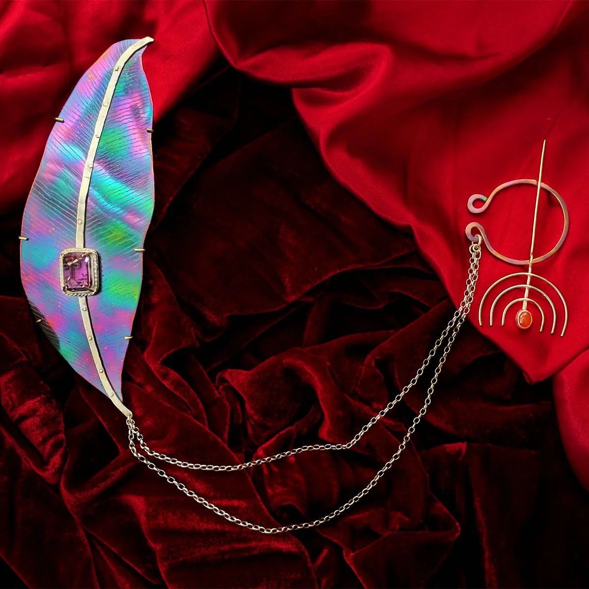 Sterling silver and titanium brooch shaped like a purple and green olive leaf with an amethyst and miniature portrait, connected by a chain to a red inverted-Menorah fibula, shown on red velvet.