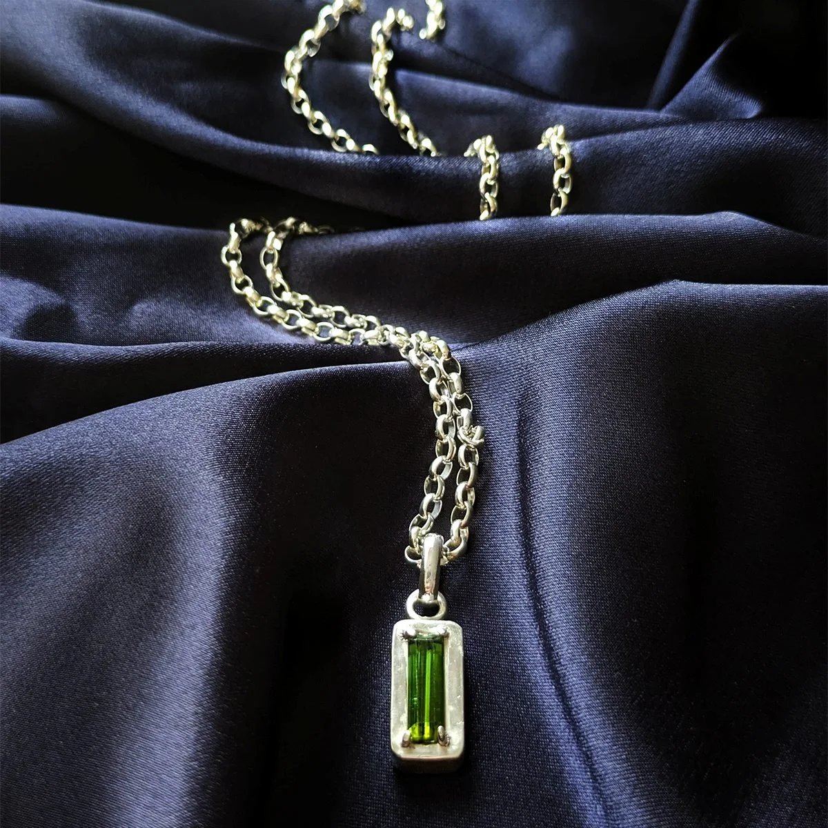 Full view of the Ohad necklace with a baguette-cut green tourmaline claw-set on a high fine silver platform, on a sterling silver Belcher chain draped over dark navy fabric.