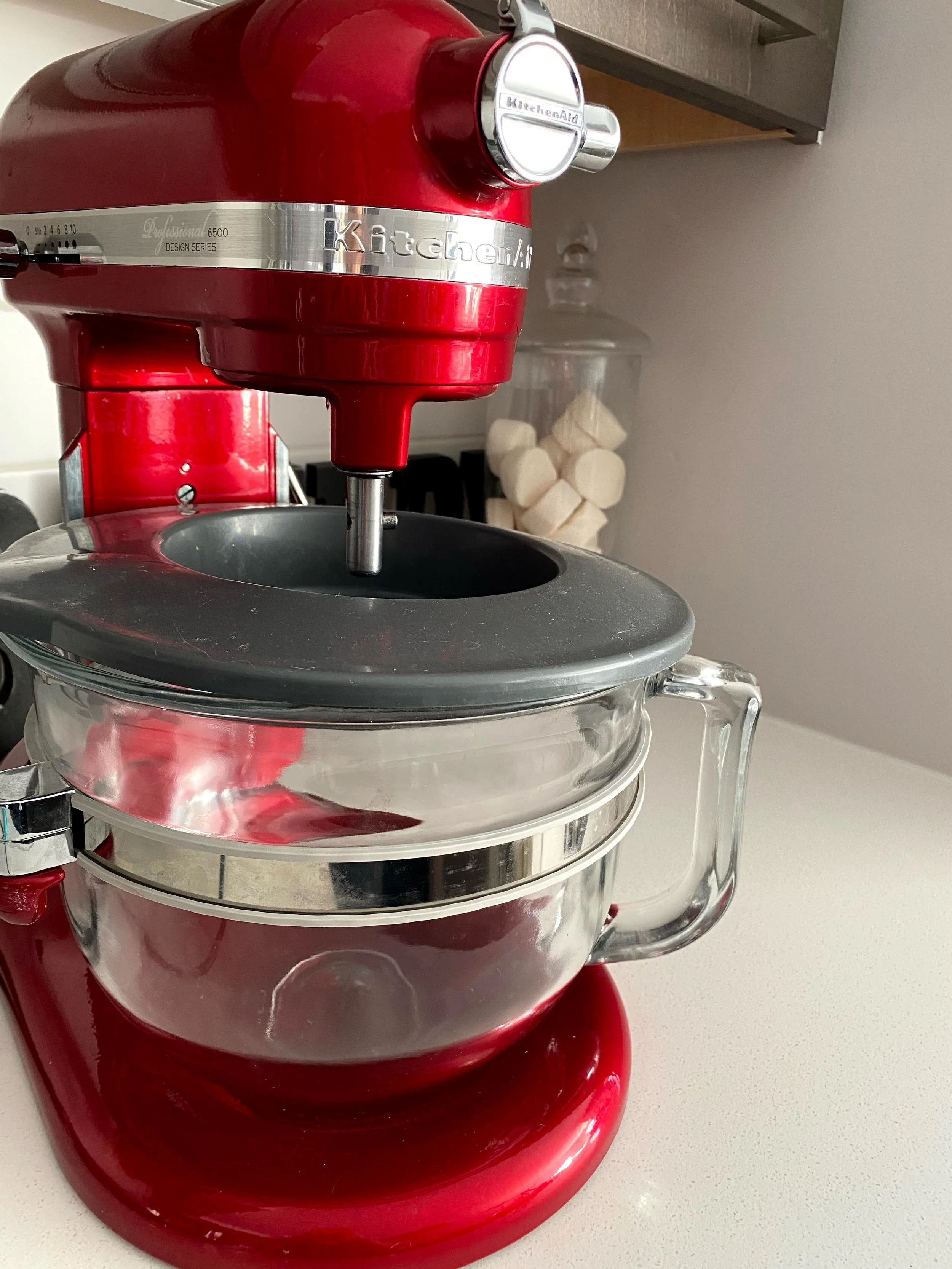 Red stand mixer with a glass bowl on a kitchen countertop, with a transparent jar of marshmallows in the background.