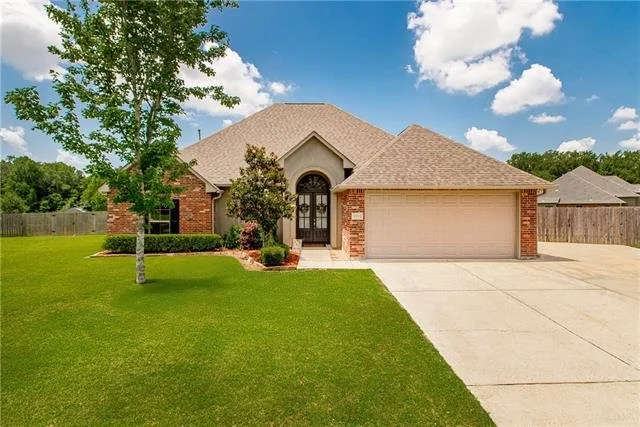 A single-story brick house with a two-car garage, a landscaped front yard with a tree and shrubs, and a concrete driveway on a bright, partly cloudy day.