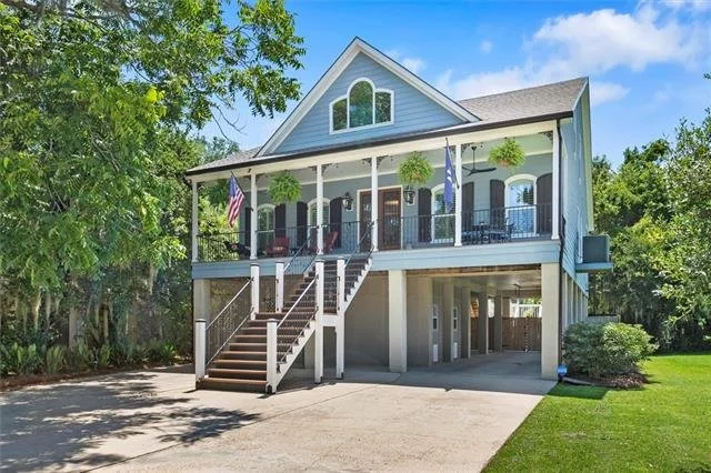 A two-story house with a large front porch, staircase, and hanging plants, surrounded by trees and greenery.