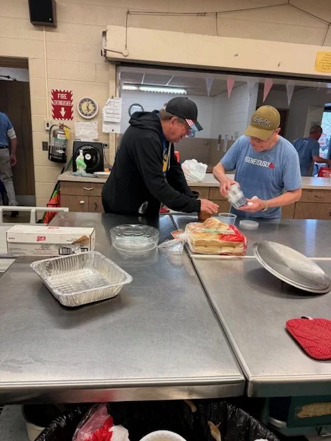 Two men preparing sandwiches at a stainless steel counter in a kitchen or cafeteria, with trays, bread, and utensils on the counter.