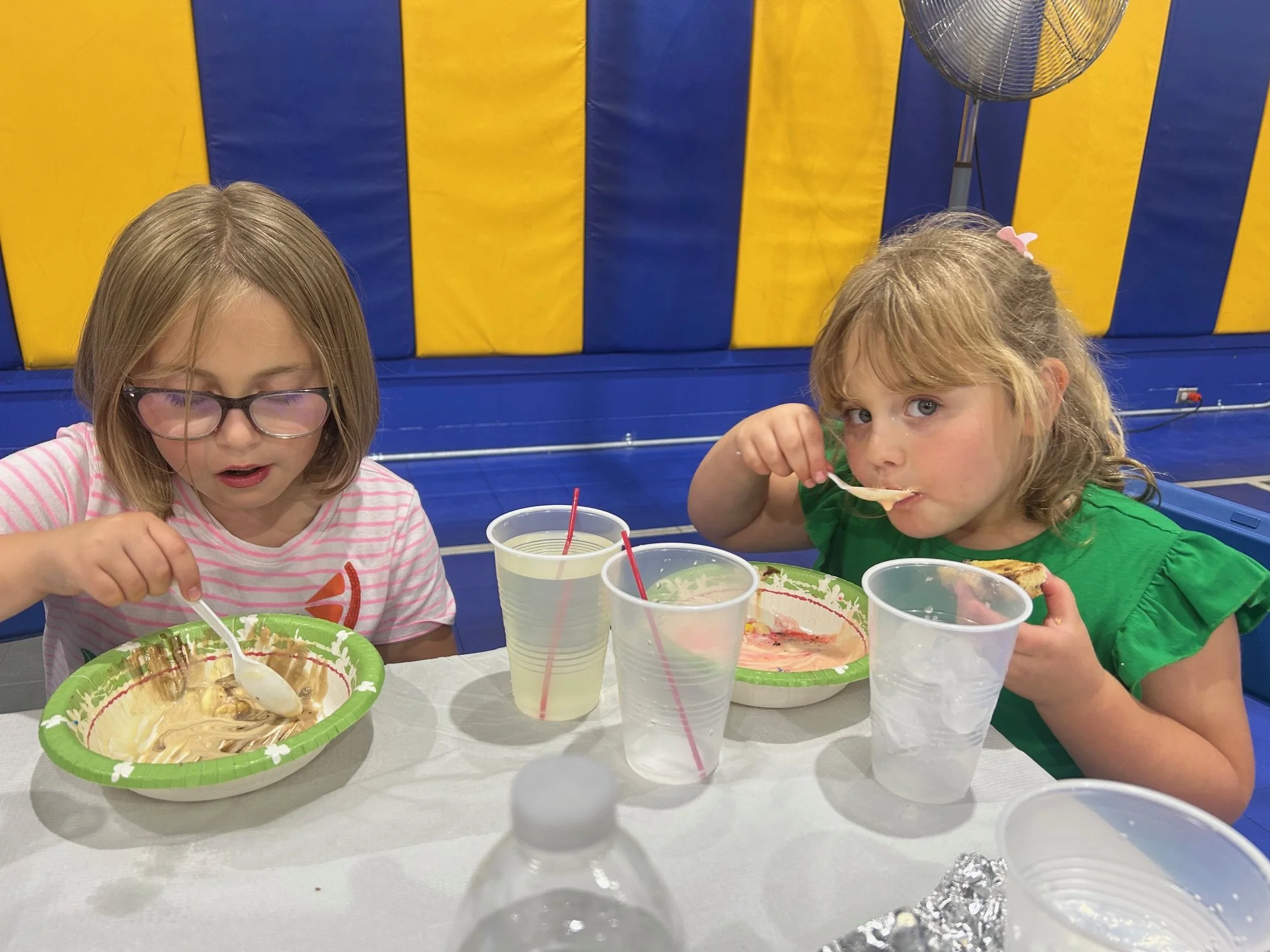 Two young girls sitting at a table eating ice cream or dessert from green paper bowls. They are surrounded by clear plastic cups with red straws, and there are some empty or partially consumed drinks on the table. The background features a blue and y