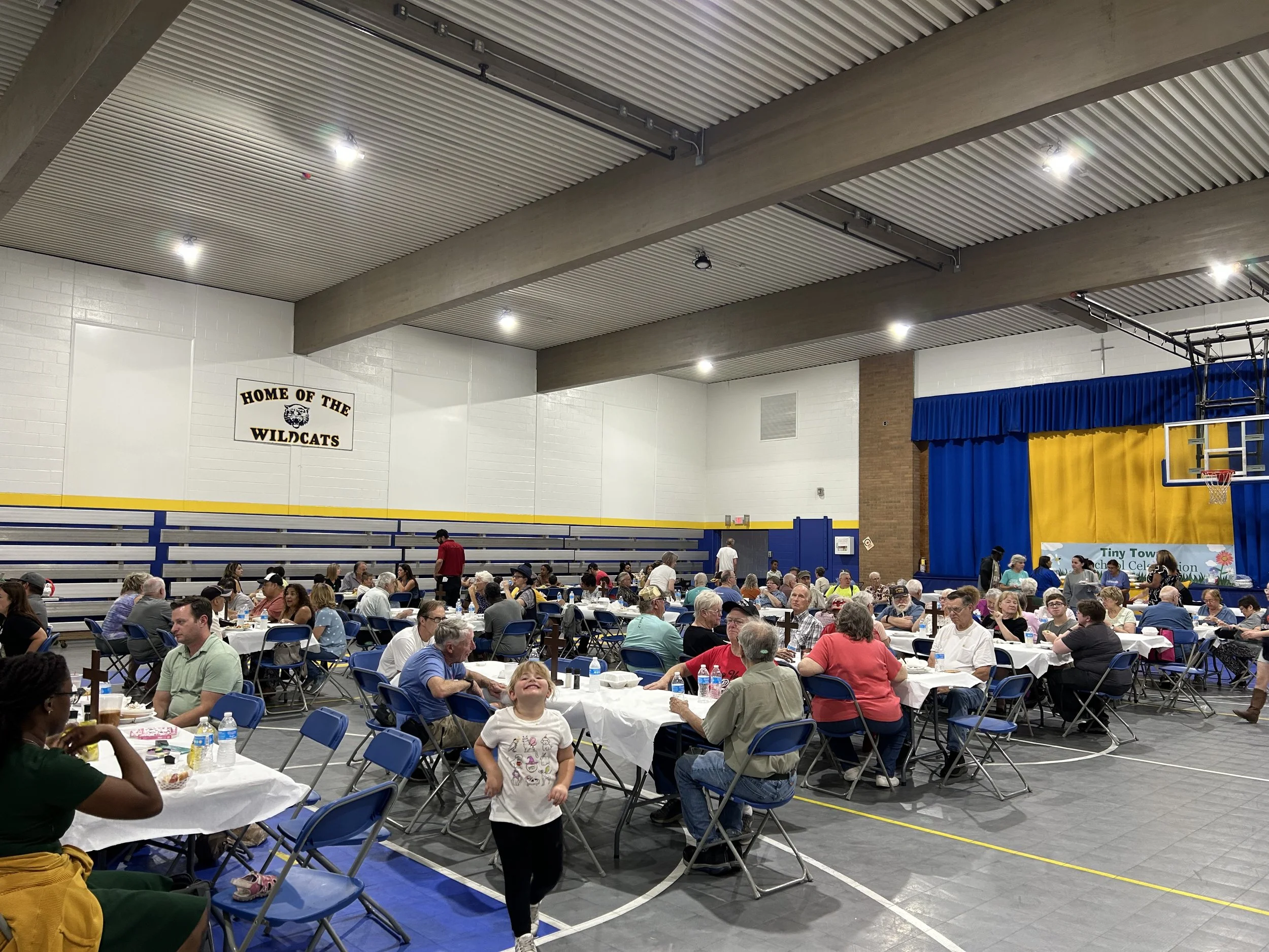A large indoor gymnasium filled with many round tables covered with white tablecloths and chairs. People of various ages are seated and standing around, eating and socializing. A young girl in the foreground is smiling at the camera. The gym has a hi
