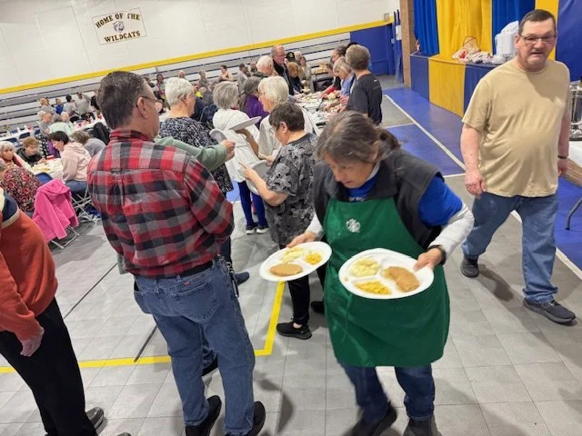 People line up in a gymnasium, where a woman in a green apron is serving food on plates, with a crowd waiting in line behind her.