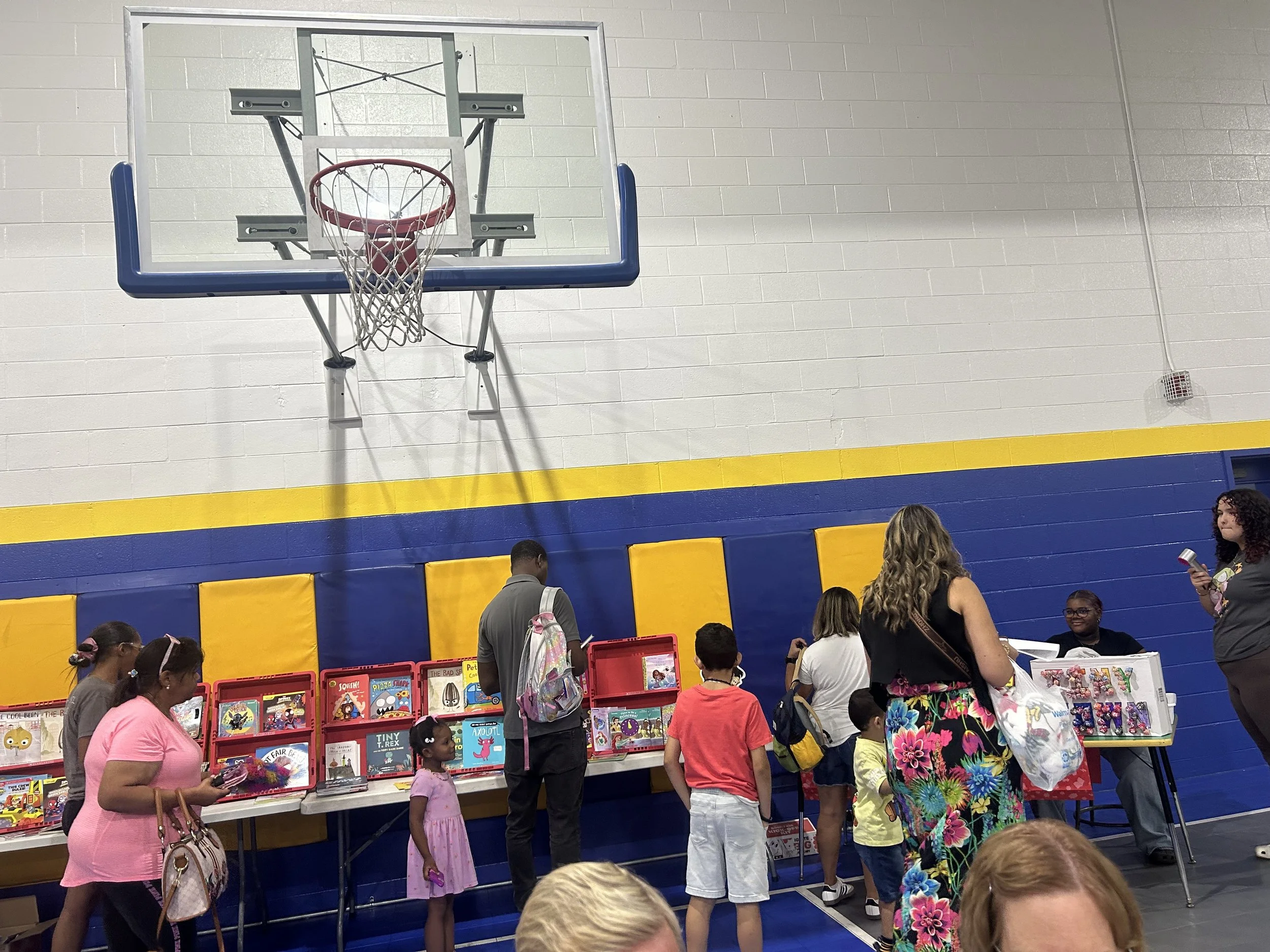 People browsing books at a book fair inside a gymnasium with a basketball hoop on the wall.