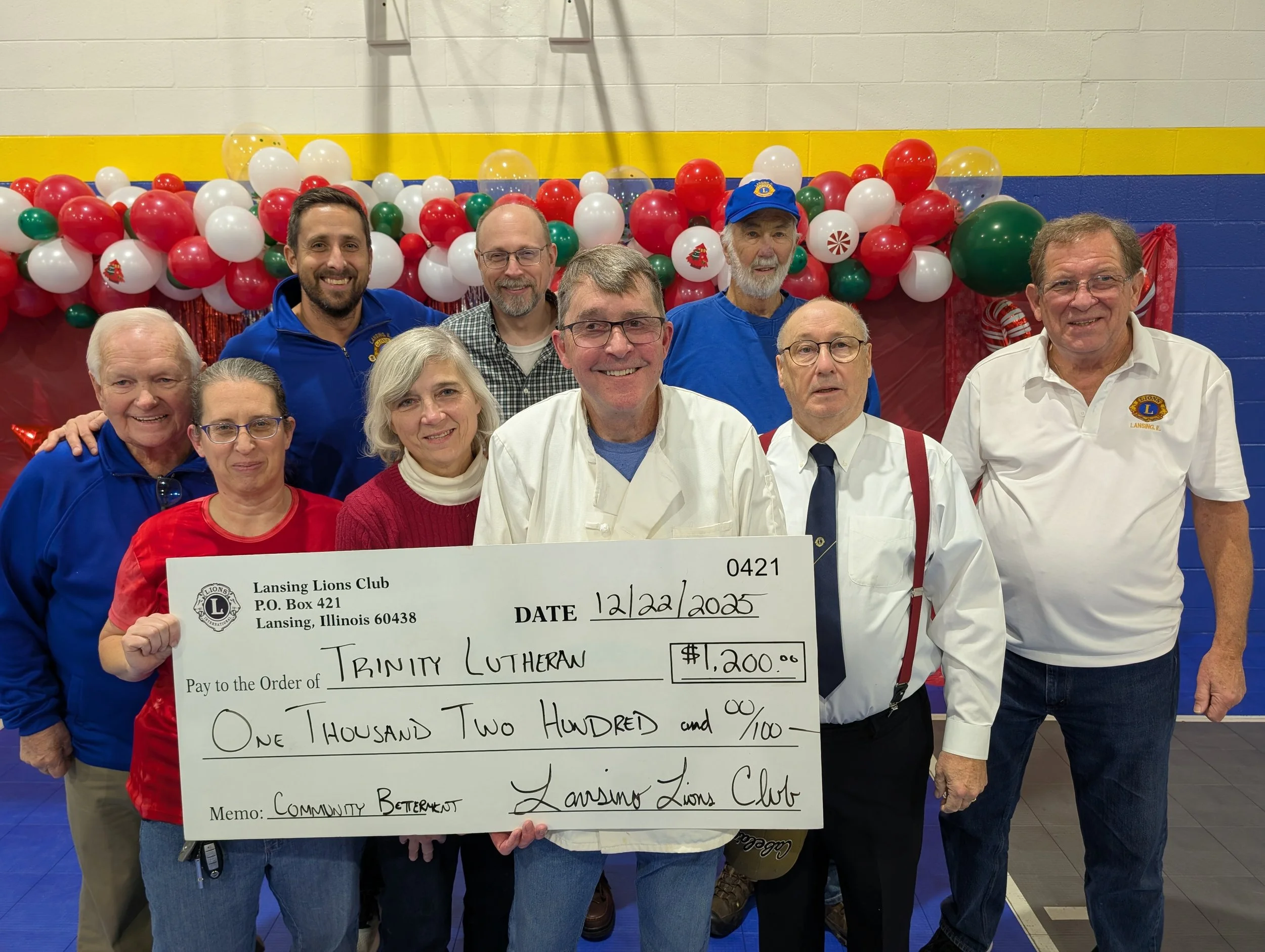 Group of nine people, seven men and two women, standing inside a gymnasium or community center, holding a large check made out to Trinity Lutheran with a date of 12/22/2025 for $1,200, presented by Lansing Lions Club. They are smiling, and behind the
