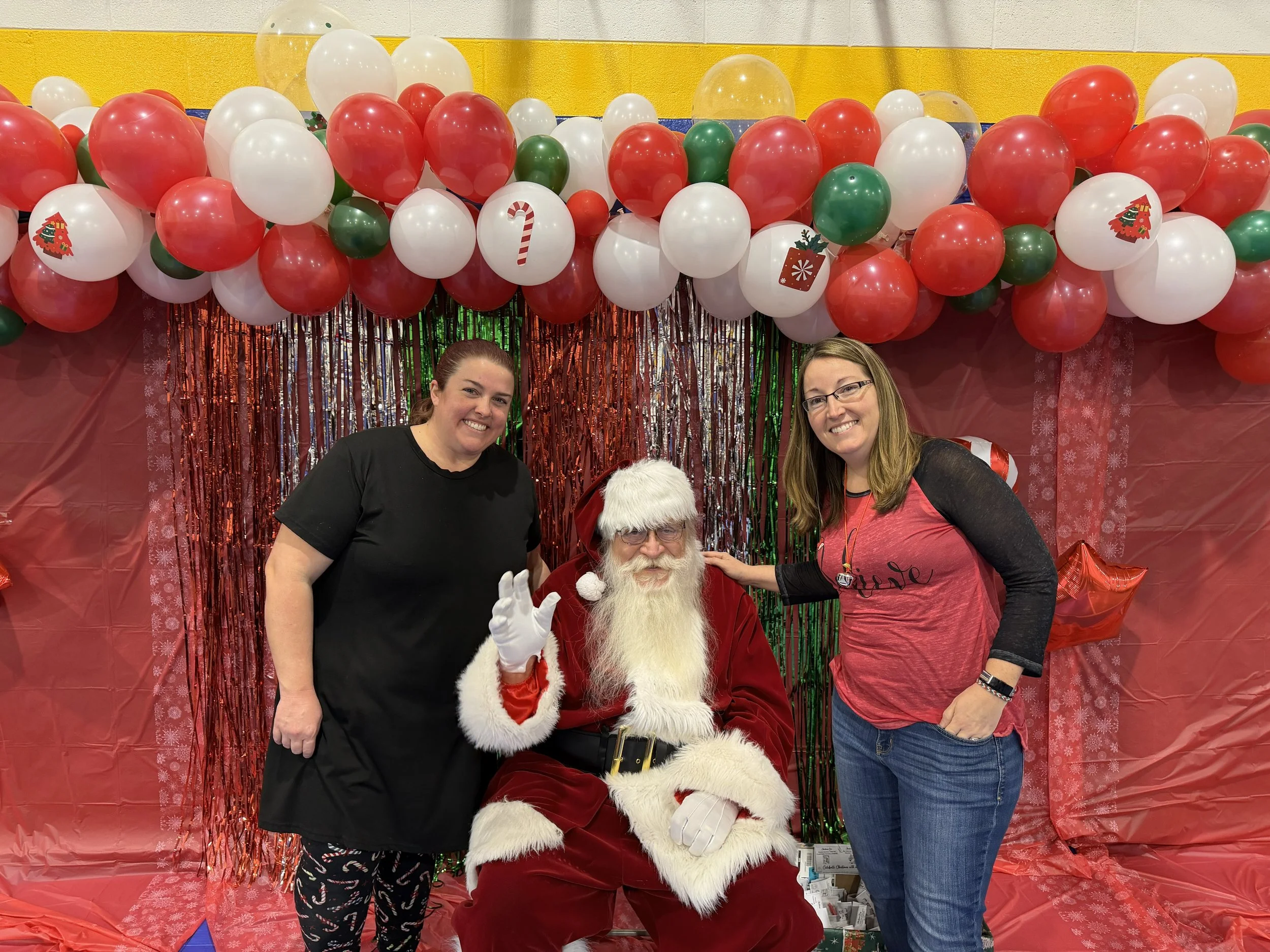 Two smiling women and a person dressed as Santa Claus sitting in front of a festive backdrop with red, white, and green balloons, Christmas decorations, and a red curtain.