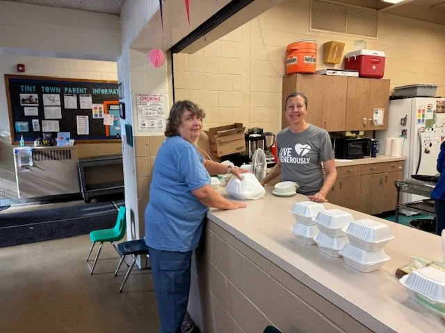 Two women smiling at a counter with food containers, in a community room or cafeteria setting.