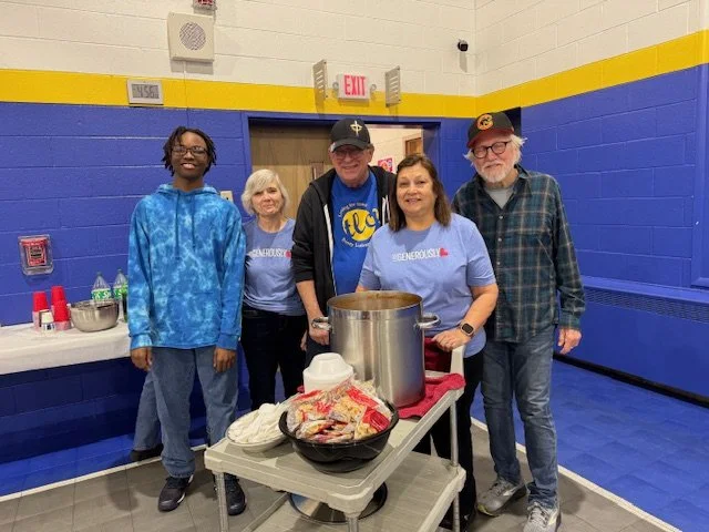 Four people standing behind a table with a large pot and food supplies in a gymnasium with blue and yellow walls, some wearing volunteer shirts.