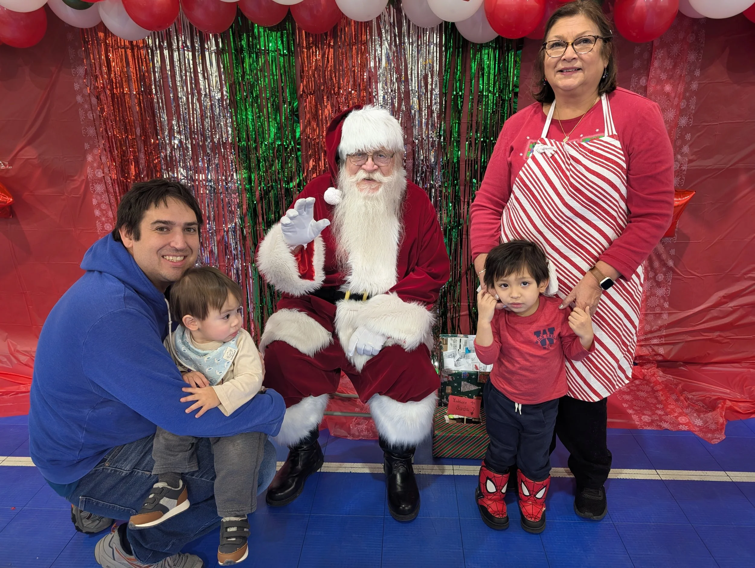 Family group photo with Santa Claus, a man in a blue hoodie, and two children, one for each side, in front of a festive holiday backdrop with balloons, tinsel, and snowflake decorations.