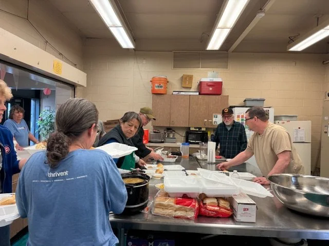 Group of people in a kitchen preparing food with various supplies and containers on the countertop.