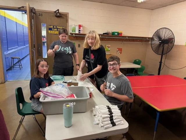 Four people in a room, two children and two adults, engaged in an activity at a table. The children are sitting, and the adults are standing, with one adult in the middle handling items on the table. The room has a red and yellow table, a large fan, 
