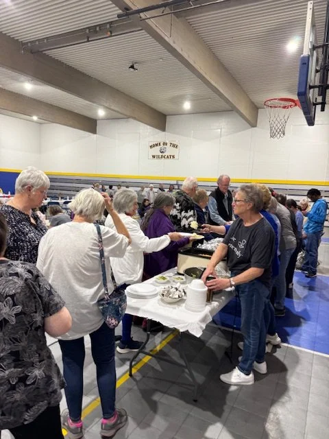 People gathered around a table with food in a gymnasium, with a sign that says "Home of the Wildcats" on the wall.