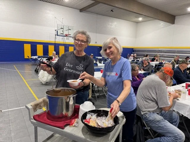 Two women serving food at a community event in a gymnasium, with tables and people in the background.