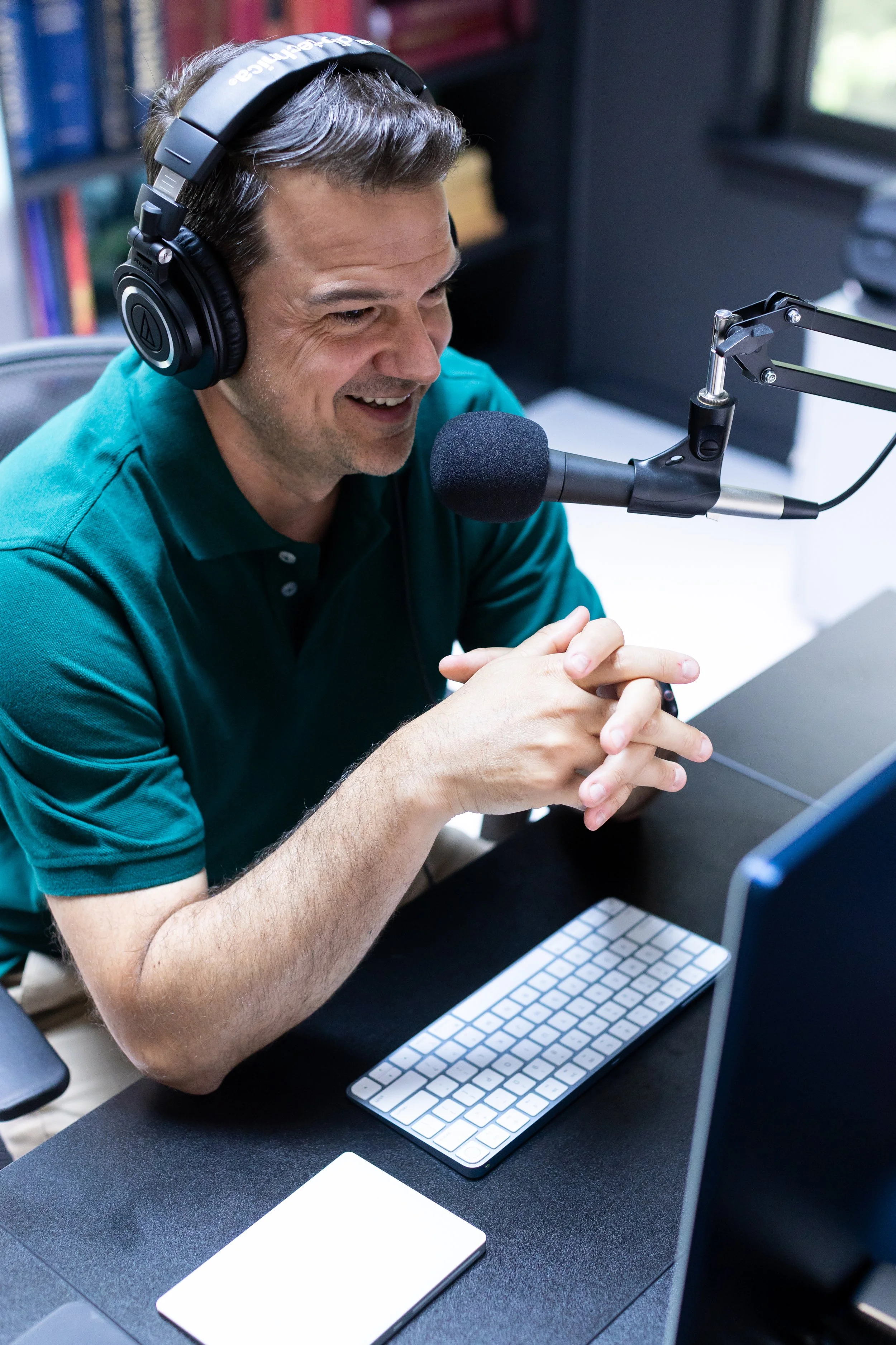 Lance Pendleton wearing headphones and a teal polo shirt, speaking into a microphone in a recording studio or radio station, with a computer monitor, keyboard, and a notepad on the desk in front of him.