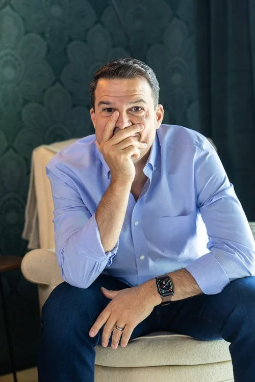 A man in a light blue dress shirt sits on a beige chair, covering his mouth with his hand, with a serious expression. He wears a smartwatch on his left wrist and a wedding band on his ring finger. The background features a dark teal patterned wall and a window with dark curtains.